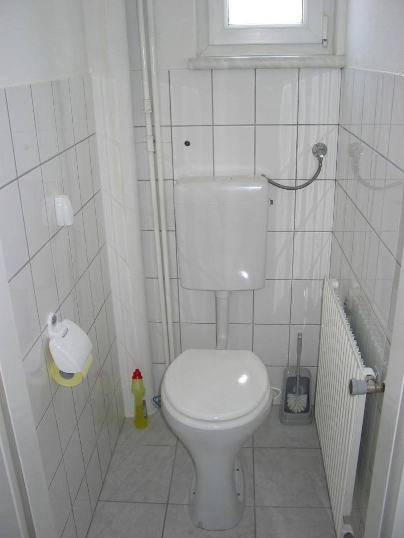 Einfache Toilette mit Fenster, weiß gefliesten Wänden und einem Heizkörper.