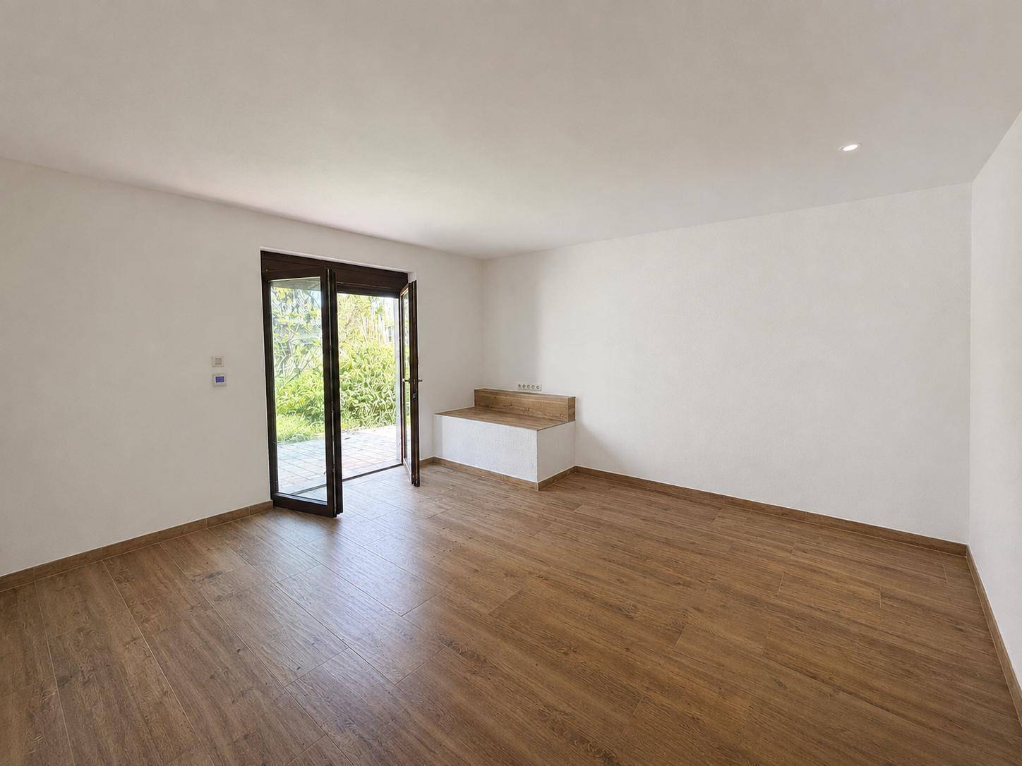Heller Raum mit Holzboden, großer Glastür und Zugang zum Außenbereich, ausgestattet mit einer Sitzbank.