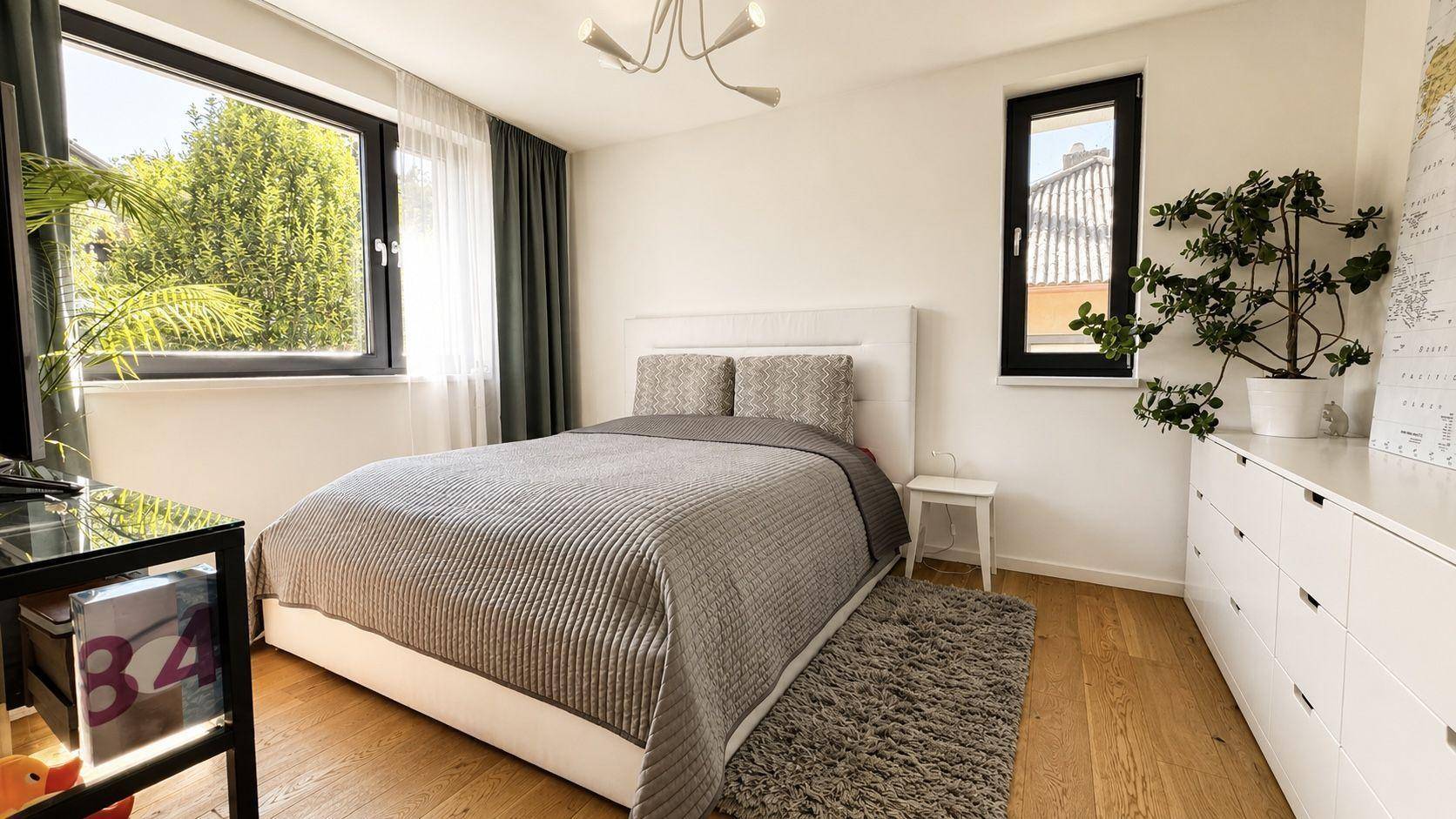 Gemütliches Schlafzimmer mit Doppelbett, weißer Kommode und zwei Fenstern für viel Tageslicht.