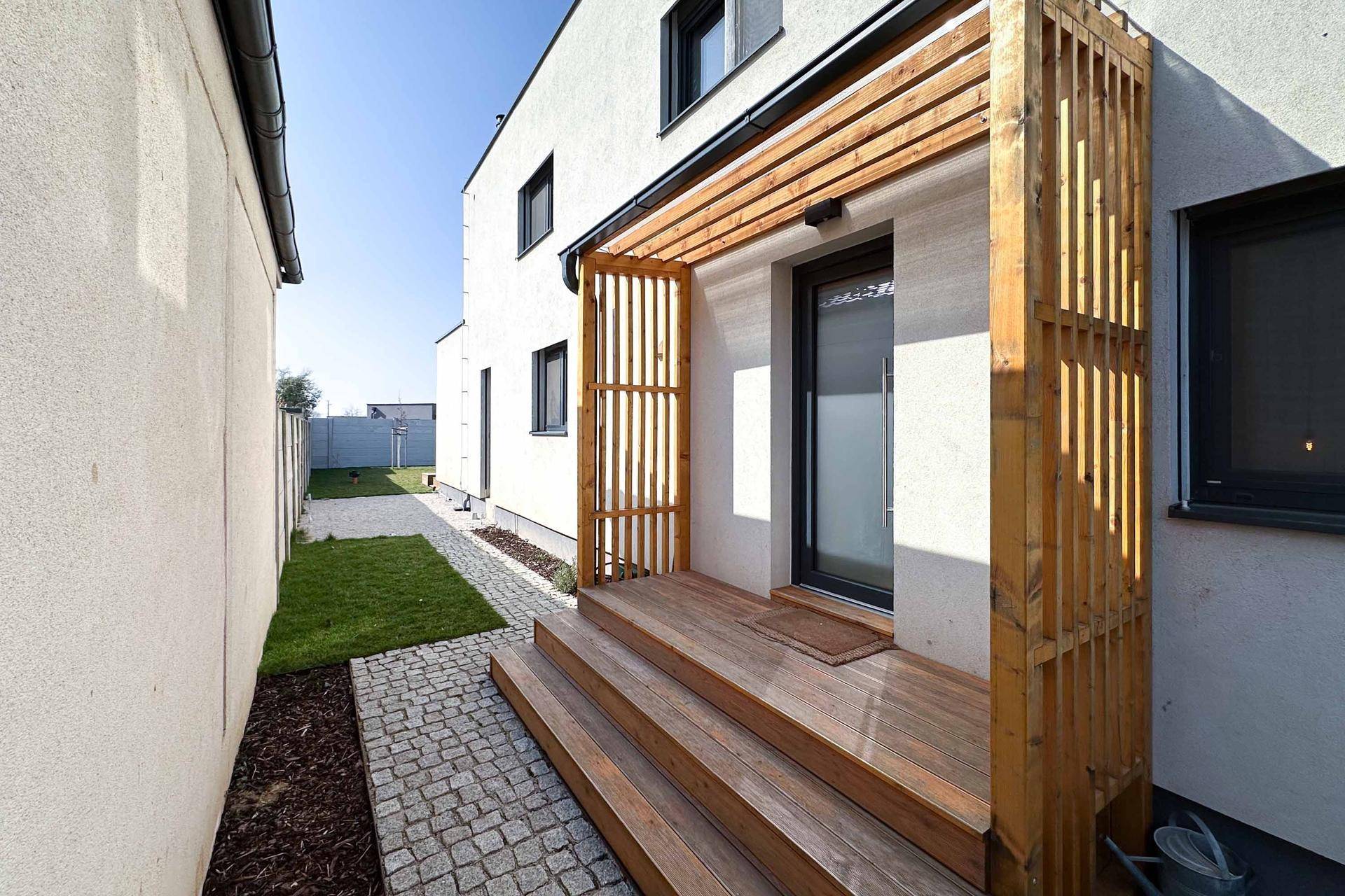 Eingangsbereich mit Holzterrasse, Pergola und gepflastertem Weg.