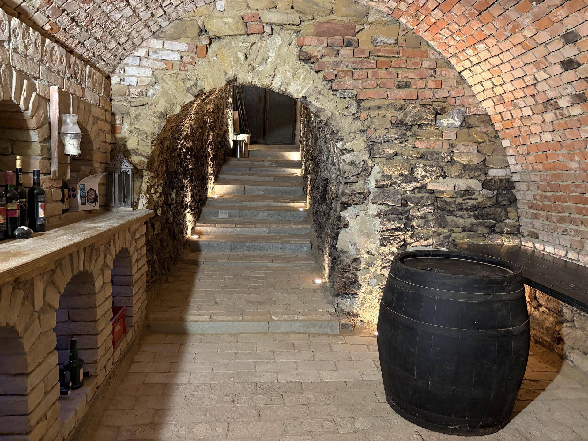 Rustikaler Weinkeller mit Ziegelwänden, Steintreppe und einem großen Holzfass als Dekoration.