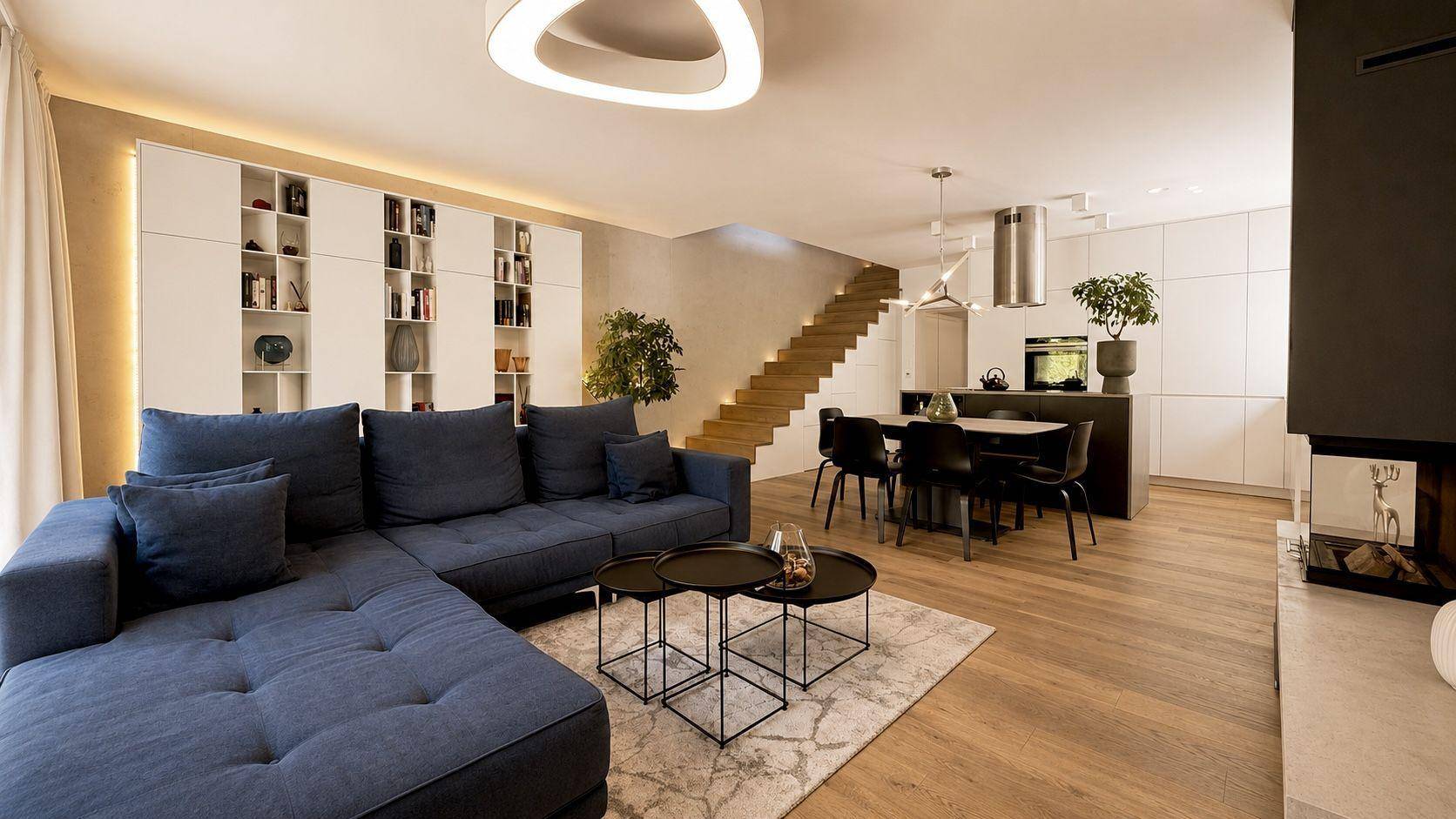 Moderner offener Wohn-Essbereich mit blauer Couch, integrierter Küche und Holztreppe zum Obergeschoss.