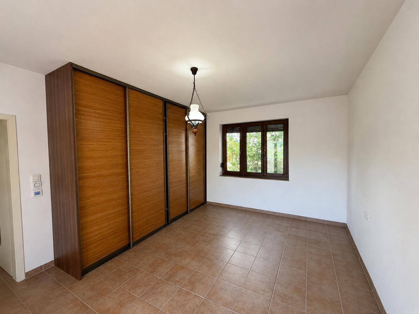 Schlafzimmer mit großem Einbauschrank, Fliesenboden und Fenster mit Blick ins Grüne.
