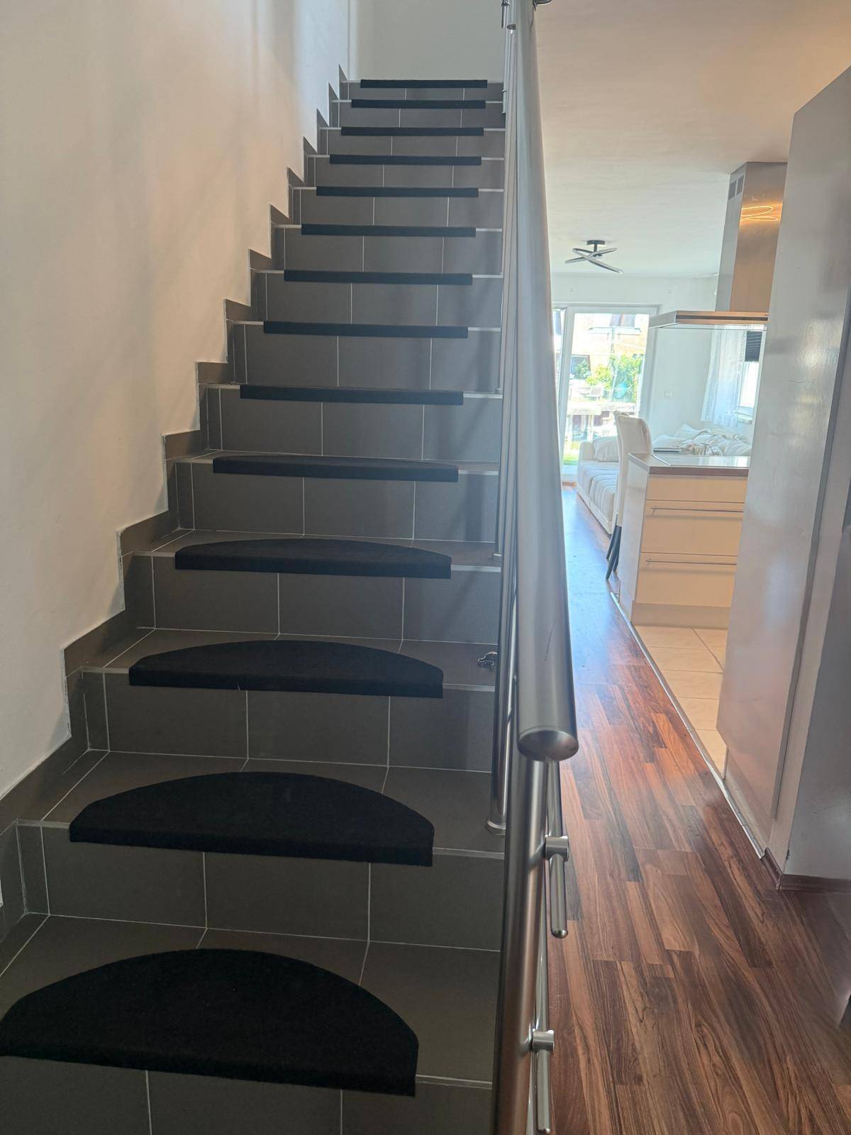 Moderne Treppe mit grauen Fliesen und Metallgeländer, angrenzend an einen hellen Wohnbereich mit Holzboden.
