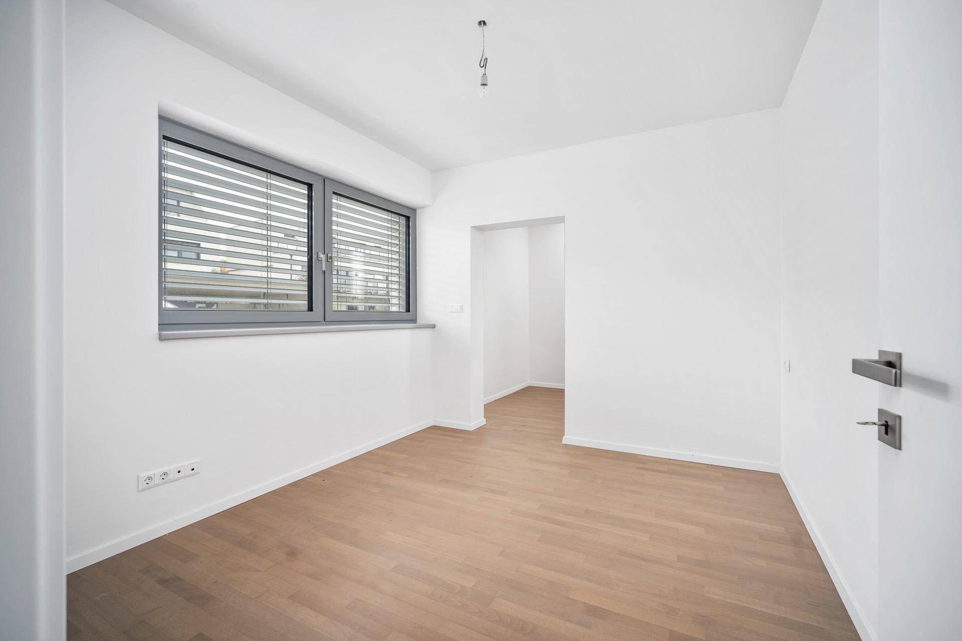 Geräumiges Zimmer mit Parkettboden, Fenster mit Jalousien und einem praktischen Nischenbereich.