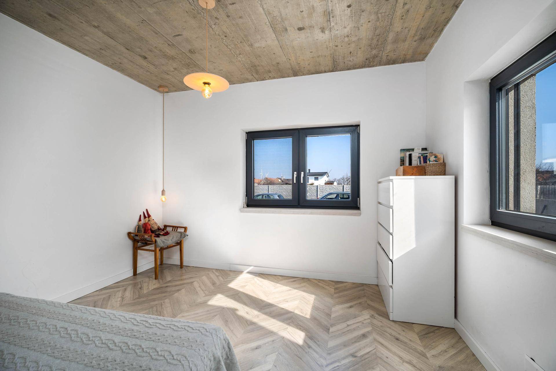 Helles Schlafzimmer mit Parkettboden, Betondecke und zwei Fenstern für Tageslicht.