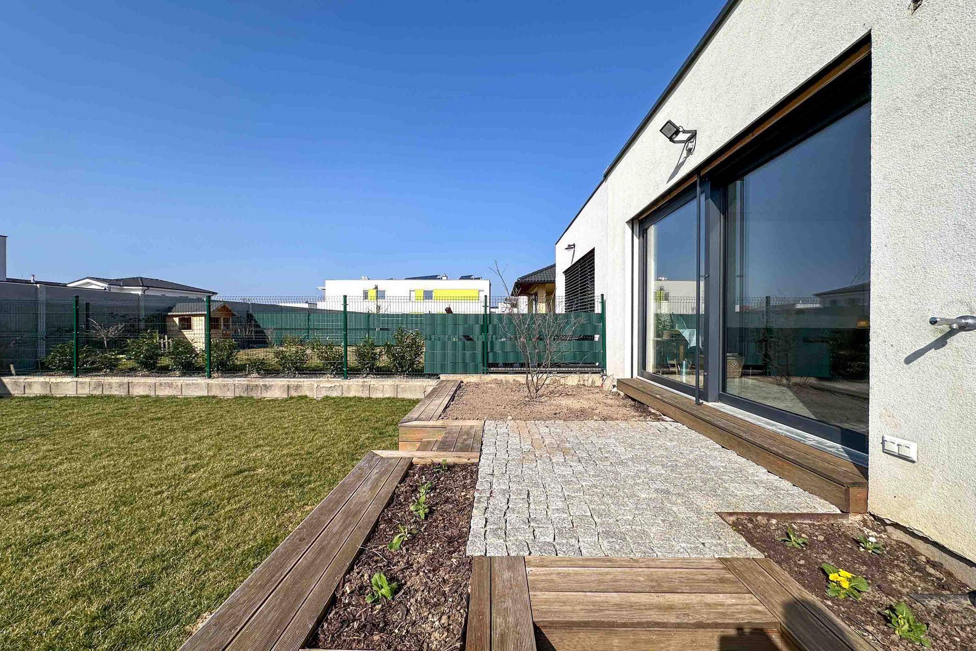 Seitlicher Gartenbereich mit Rasen, gepflasterter Terrasse und erhöhten Holzbeeten.