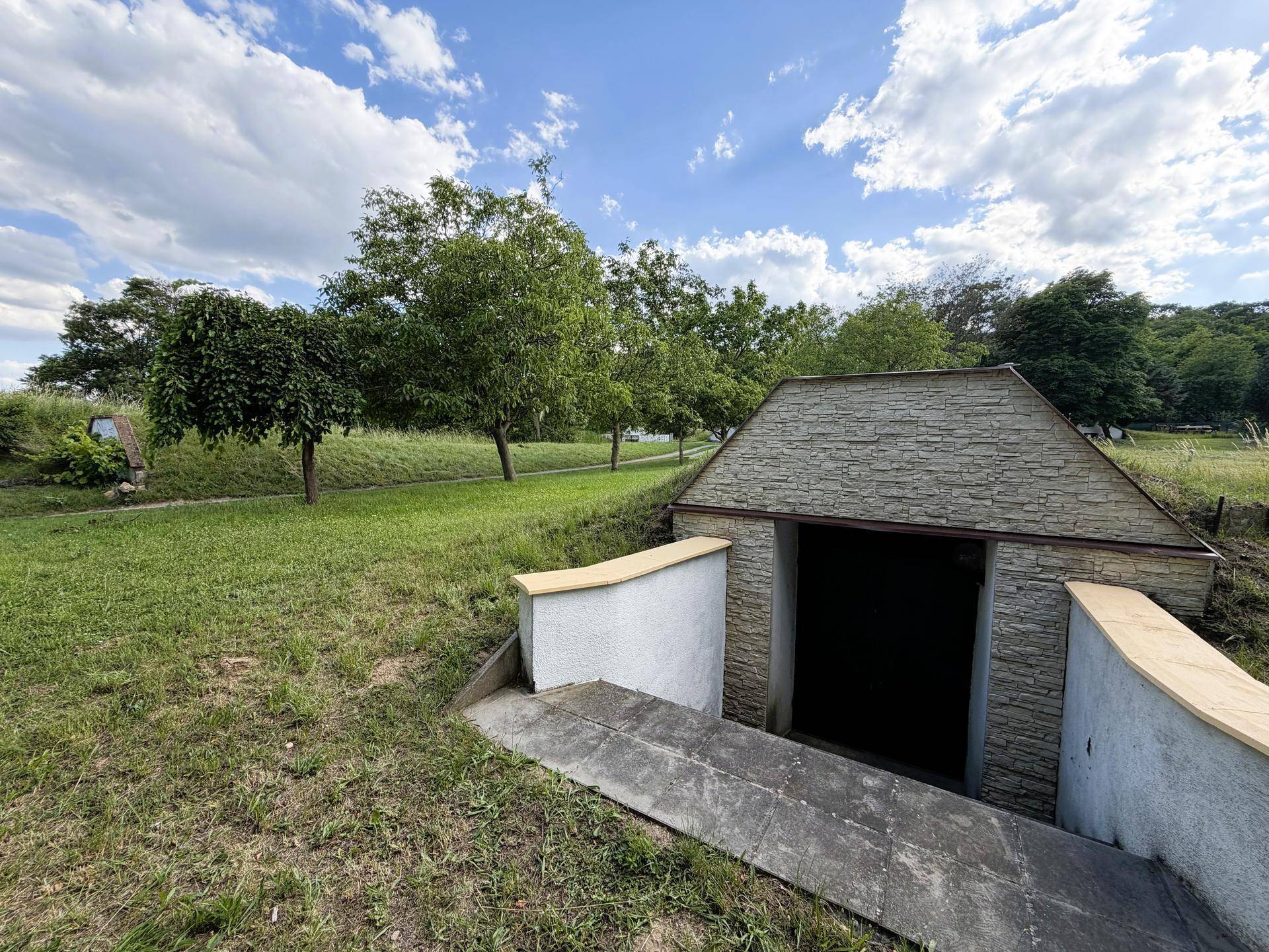 Eingang zu einem Erdkeller oder Bunker, eingebettet in eine grüne Hügellandschaft mit Bäumen.