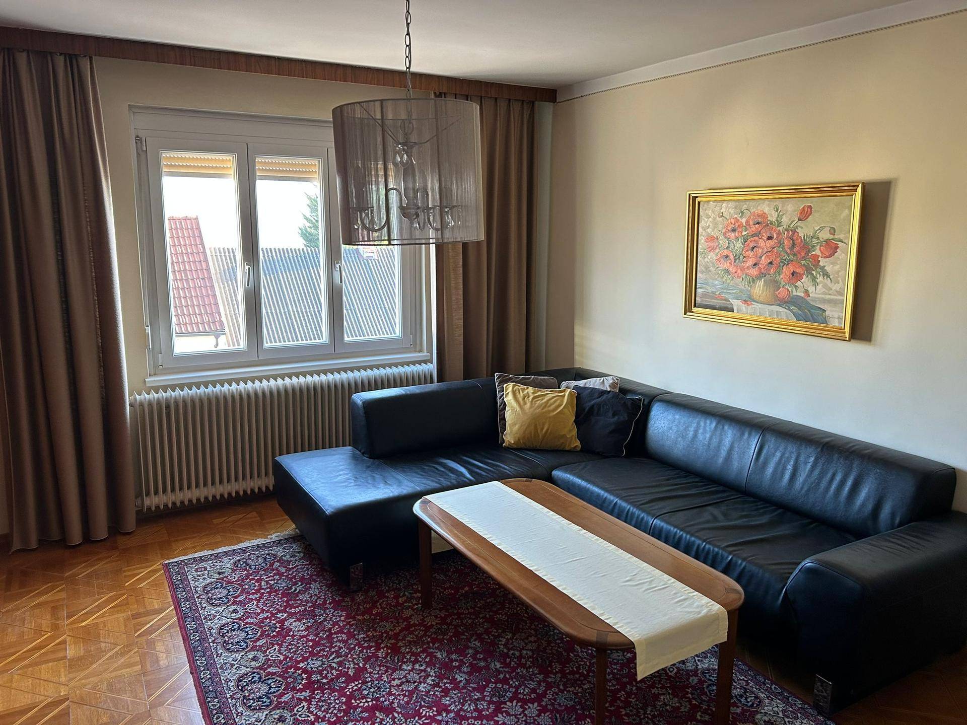 Wohnzimmer mit modernem Ecksofa, Parkettboden und großem Fenster mit Vorhängen.