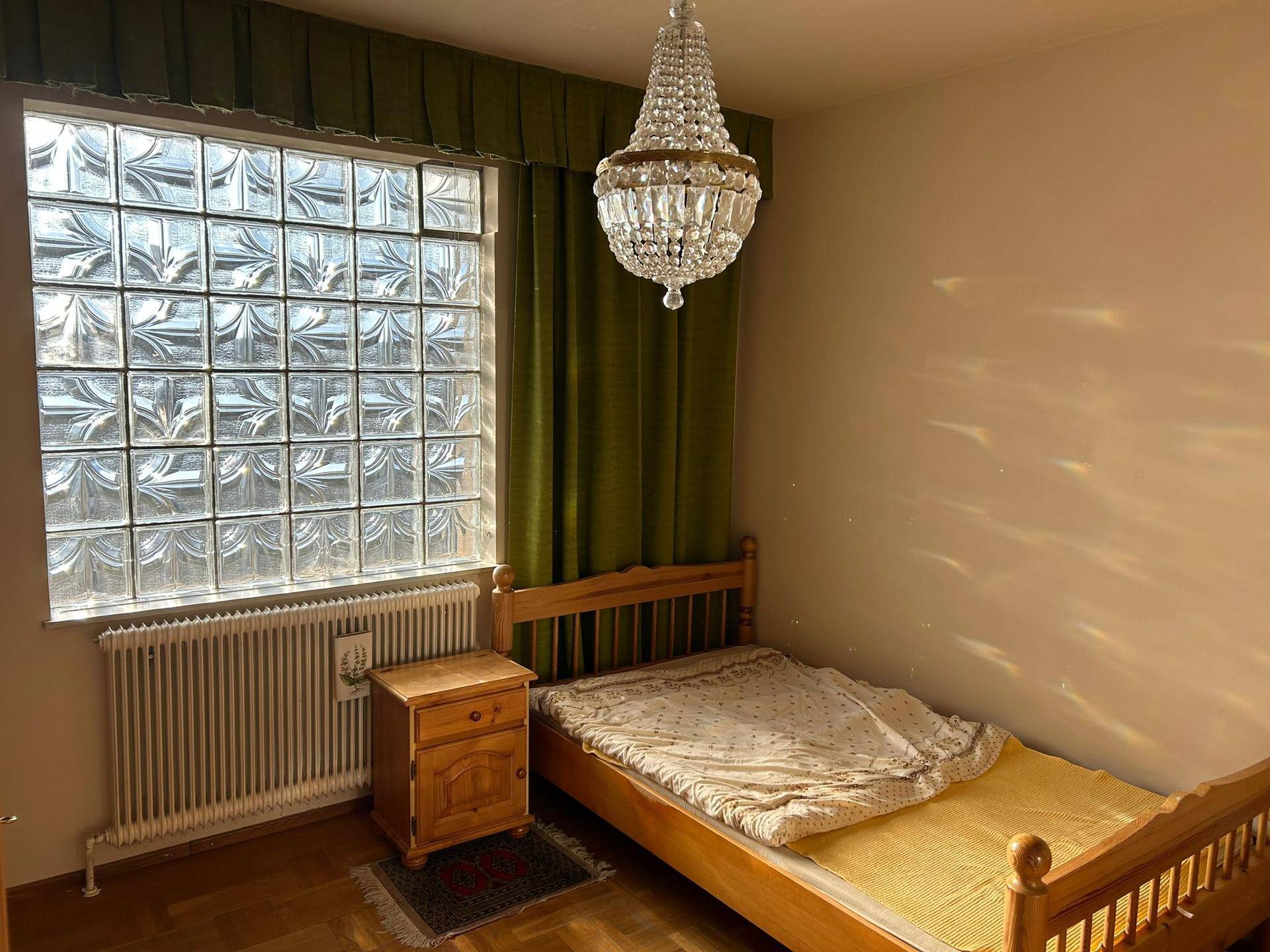Schlafzimmer mit Einzelbett, Kristallleuchter und Fenster aus Glasbausteinen.