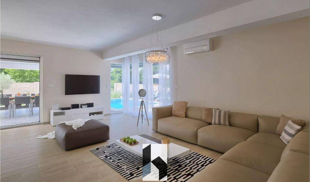 Helles Wohnzimmer mit beigem Sofa, Fernseher und Zugang zur Poolterrasse.