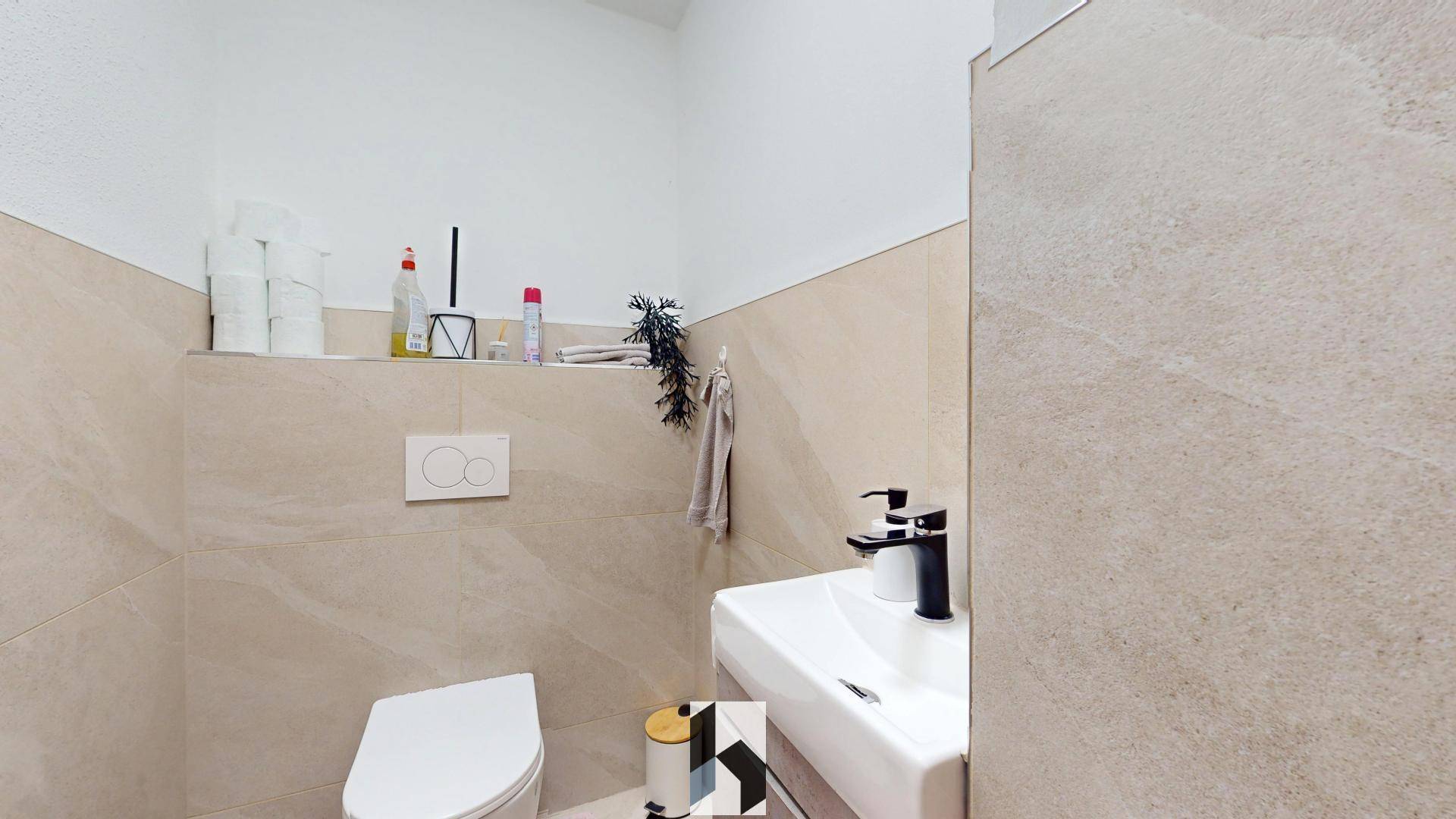 Gäste-WC mit Wand-WC, kleinem Waschbecken und beigefarbenen Fliesen an den Wänden.