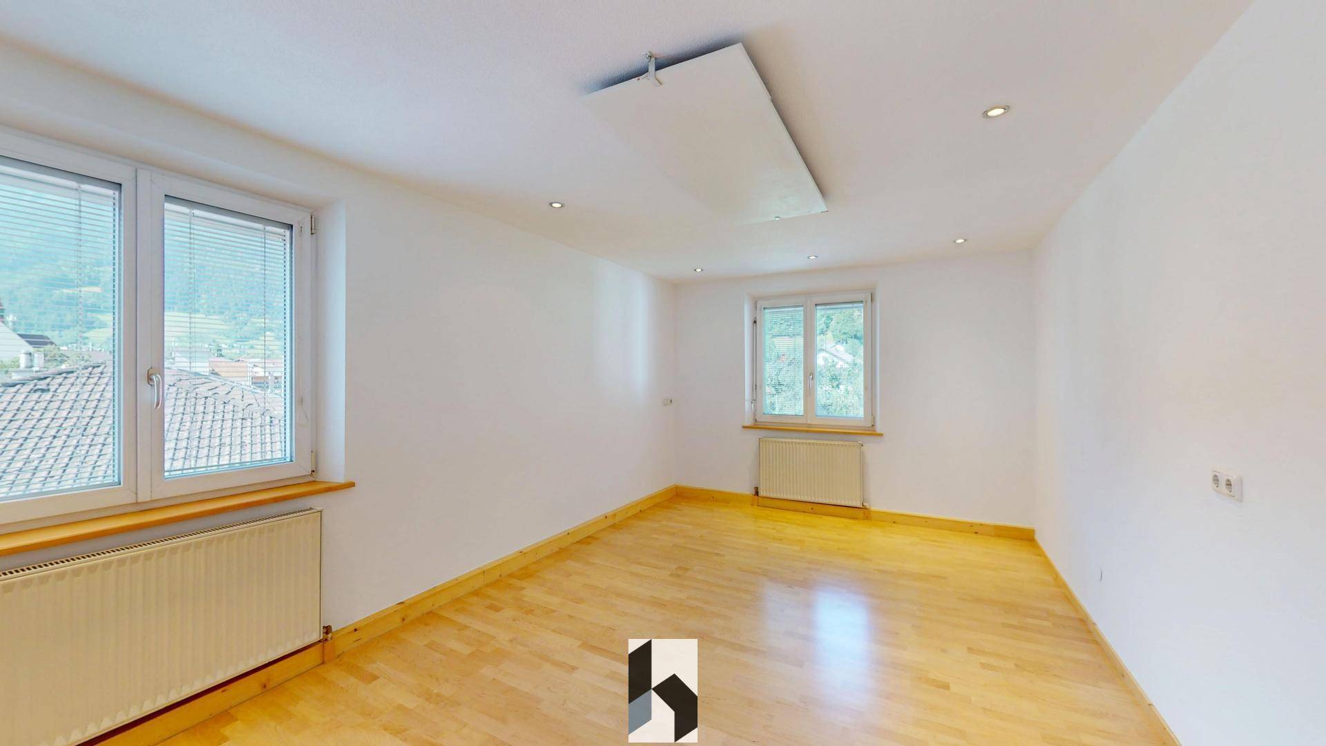 Helles Zimmer mit Holzboden, weißen Wänden und zwei Fenstern mit Blick auf die Umgebung.