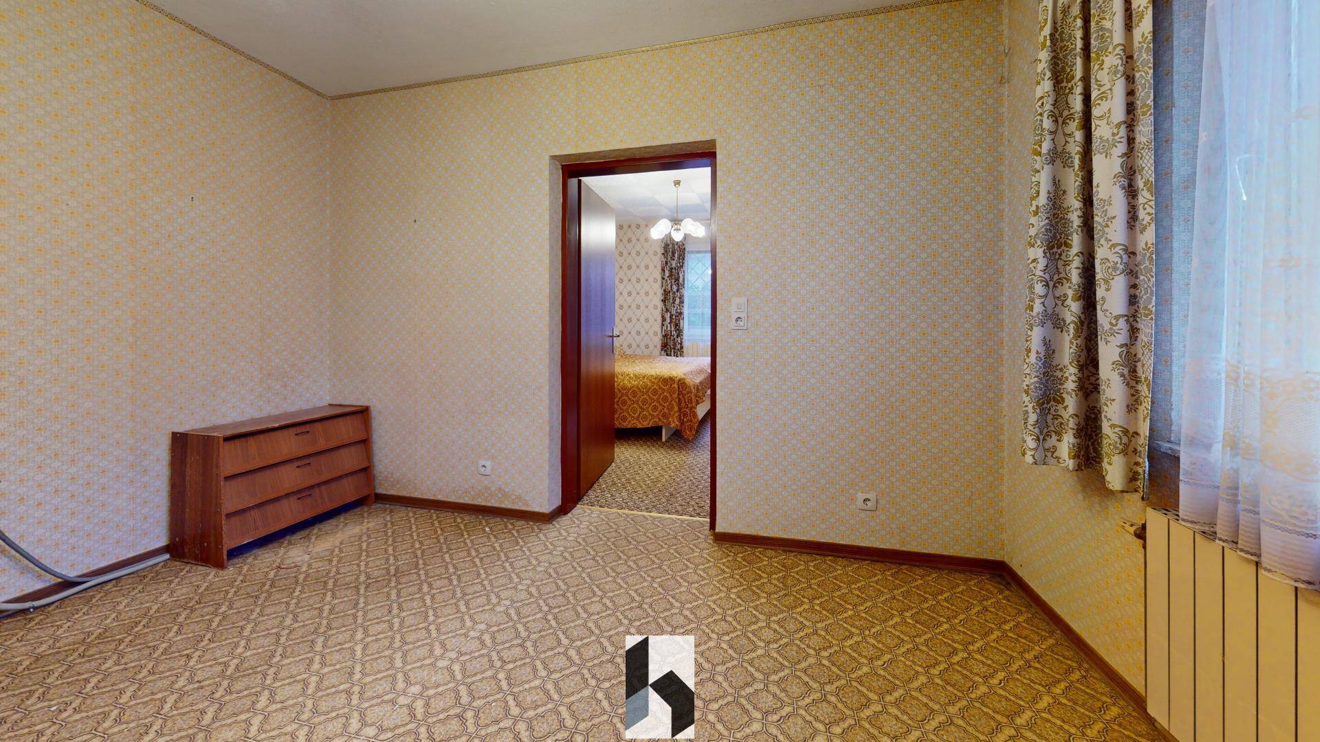 Zimmer mit gemusterter Tapete und Teppichboden, das zu einem weiteren Schlafzimmer führt.