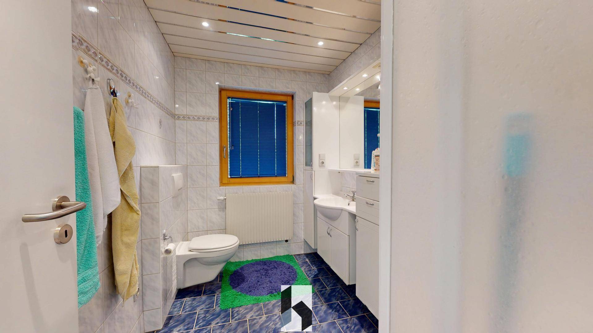 Badezimmer mit blauen Fliesen, WC, Waschbecken und einem Fenster mit Jalousien.