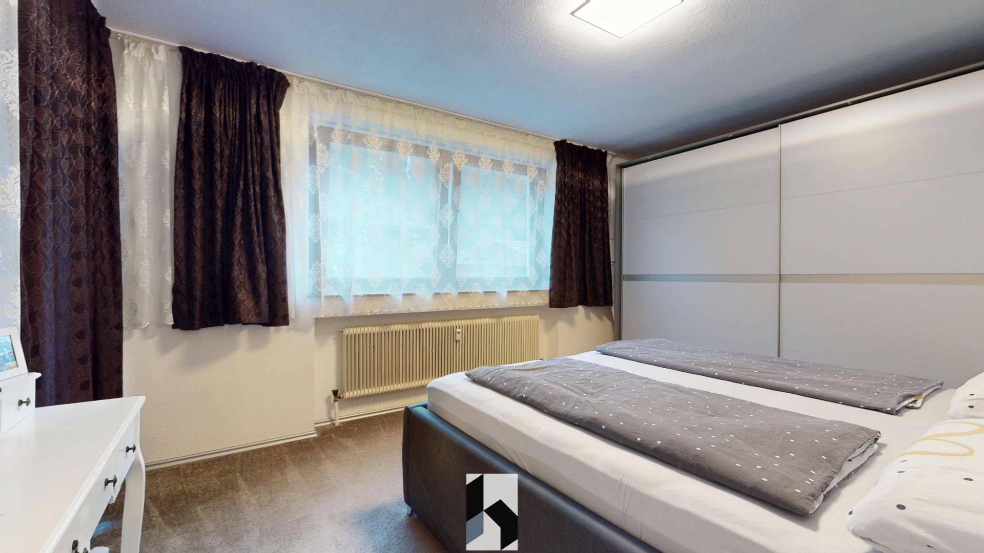 Schlafzimmer mit Doppelbett, großem weißen Schiebetürenschrank und Fenster mit Vorhängen.