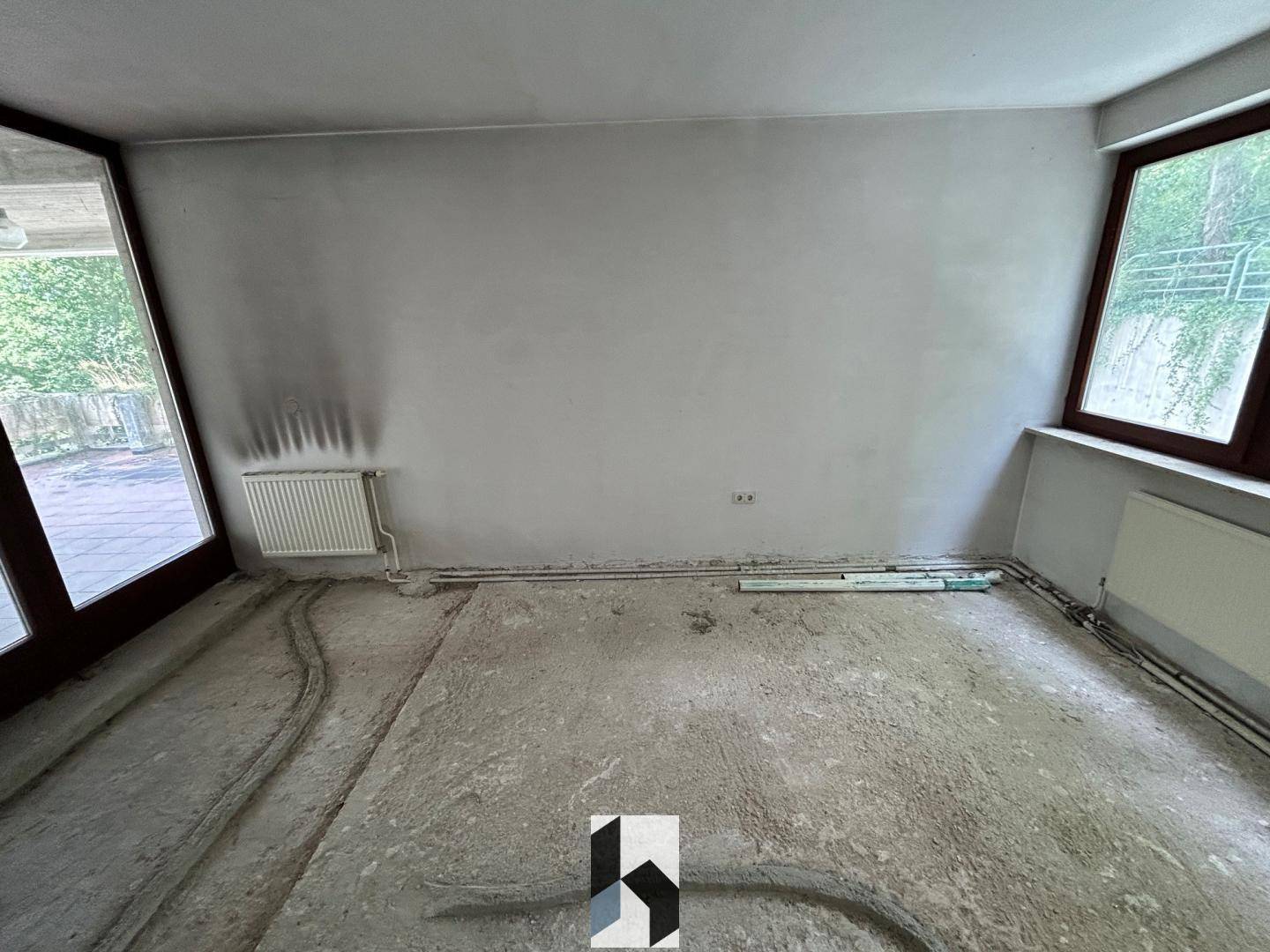 Renovierungsbedürftiger Raum mit Betonboden, weißen Wänden und großen Fenstern, bereit für den Ausbau.