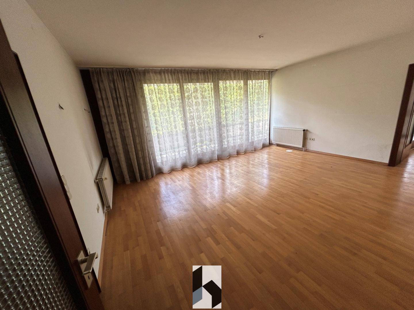Leerer Raum mit Parkettboden und großen Fenstern, die mit Vorhängen versehen sind.