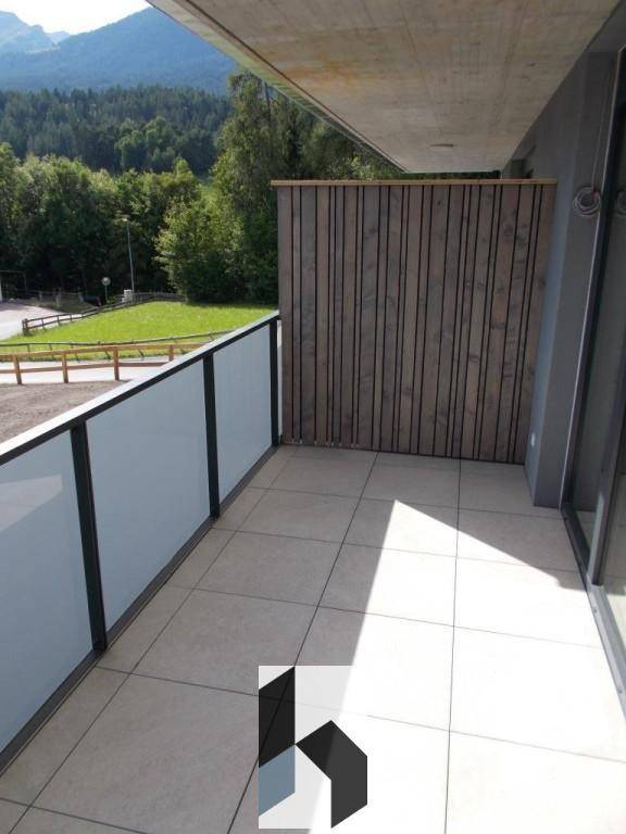 Sonniger Balkon mit Fliesenboden, Glasgeländer und schöner Aussicht auf die Berge und Natur.