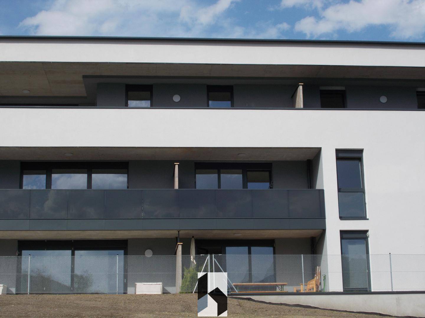 Moderne Gebäudefassade mit mehreren Balkonen und großen Fenstern in einer grünen Umgebung.
