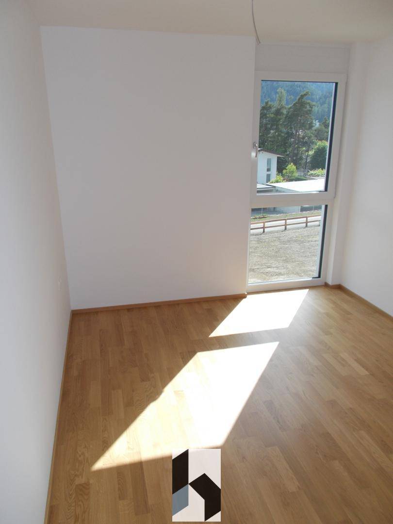 Helles Zimmer mit Holzboden und großem Fenster, das viel Tageslicht und eine grüne Aussicht bietet.