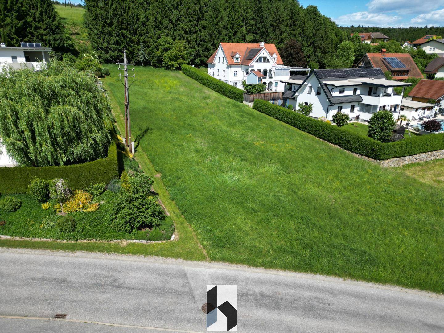 Luftaufnahme eines abfallenden, grünen Grundstücks mit Straße im Vordergrund und Wald im Hintergrund.