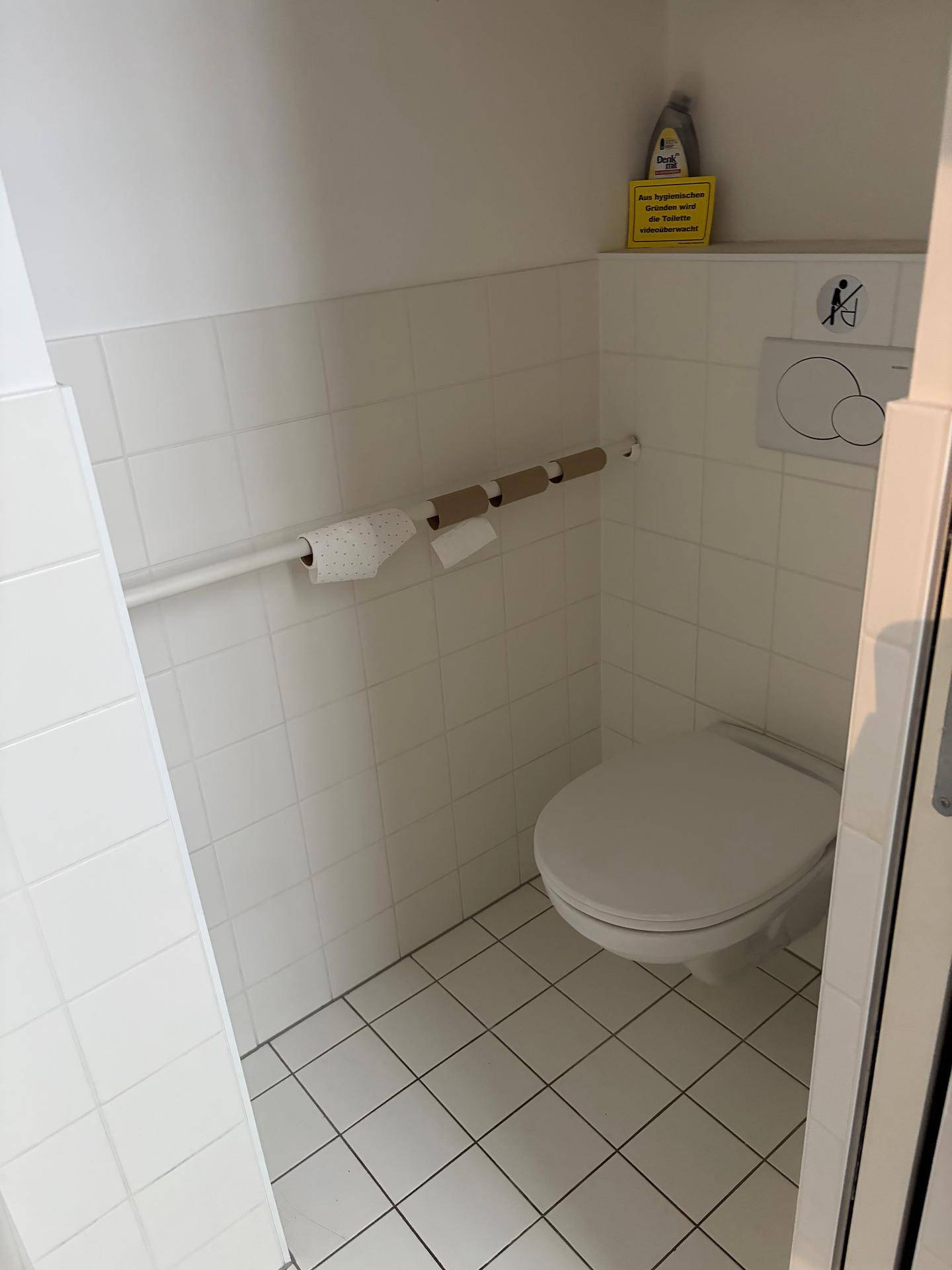 Separates WC mit Wand-WC, weißen Fliesen und Haltegriffen für Barrierefreiheit.