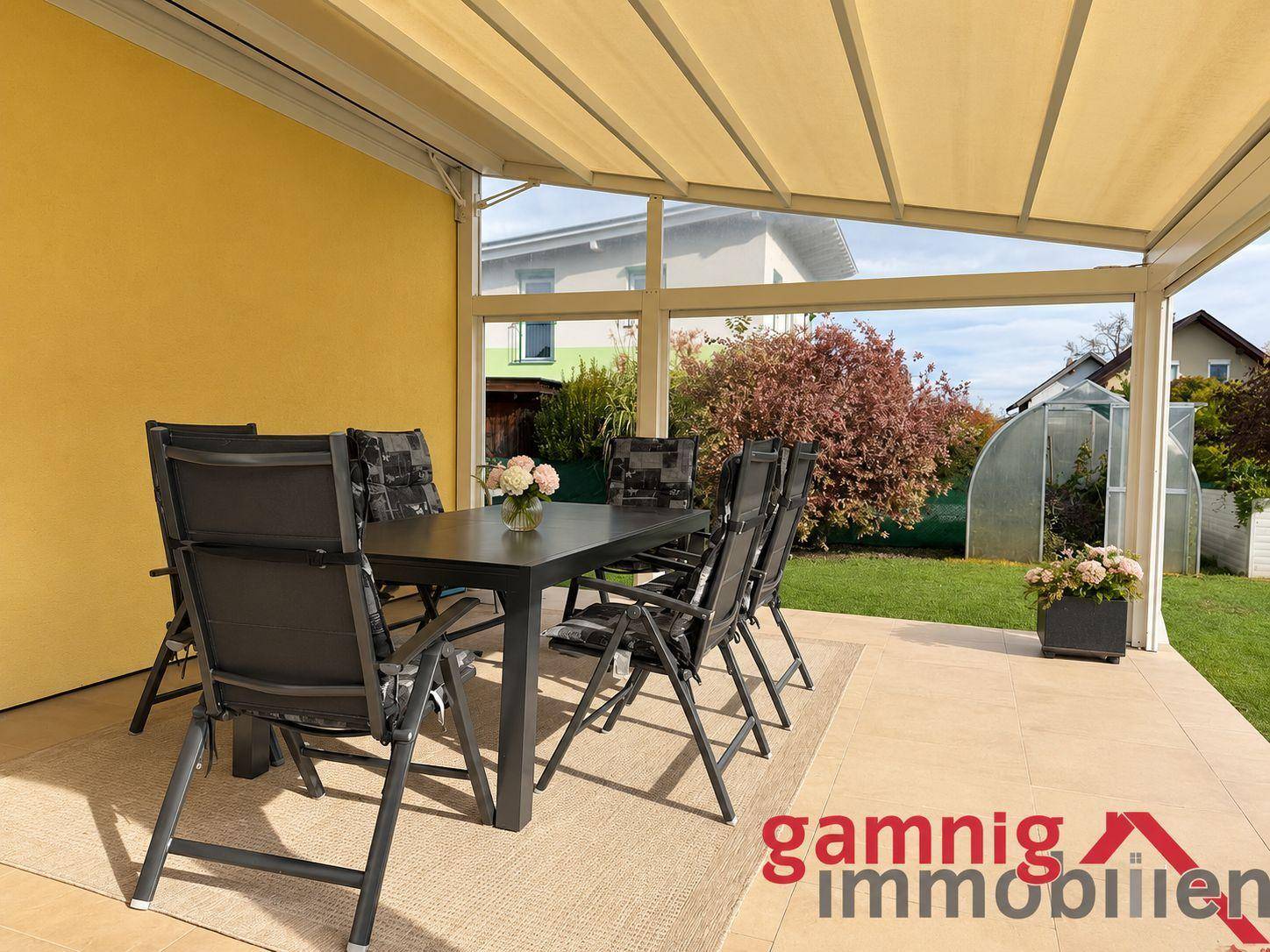 Gemütliche, überdachte Terrasse mit Esstisch und Stühlen, umgeben von Grün.