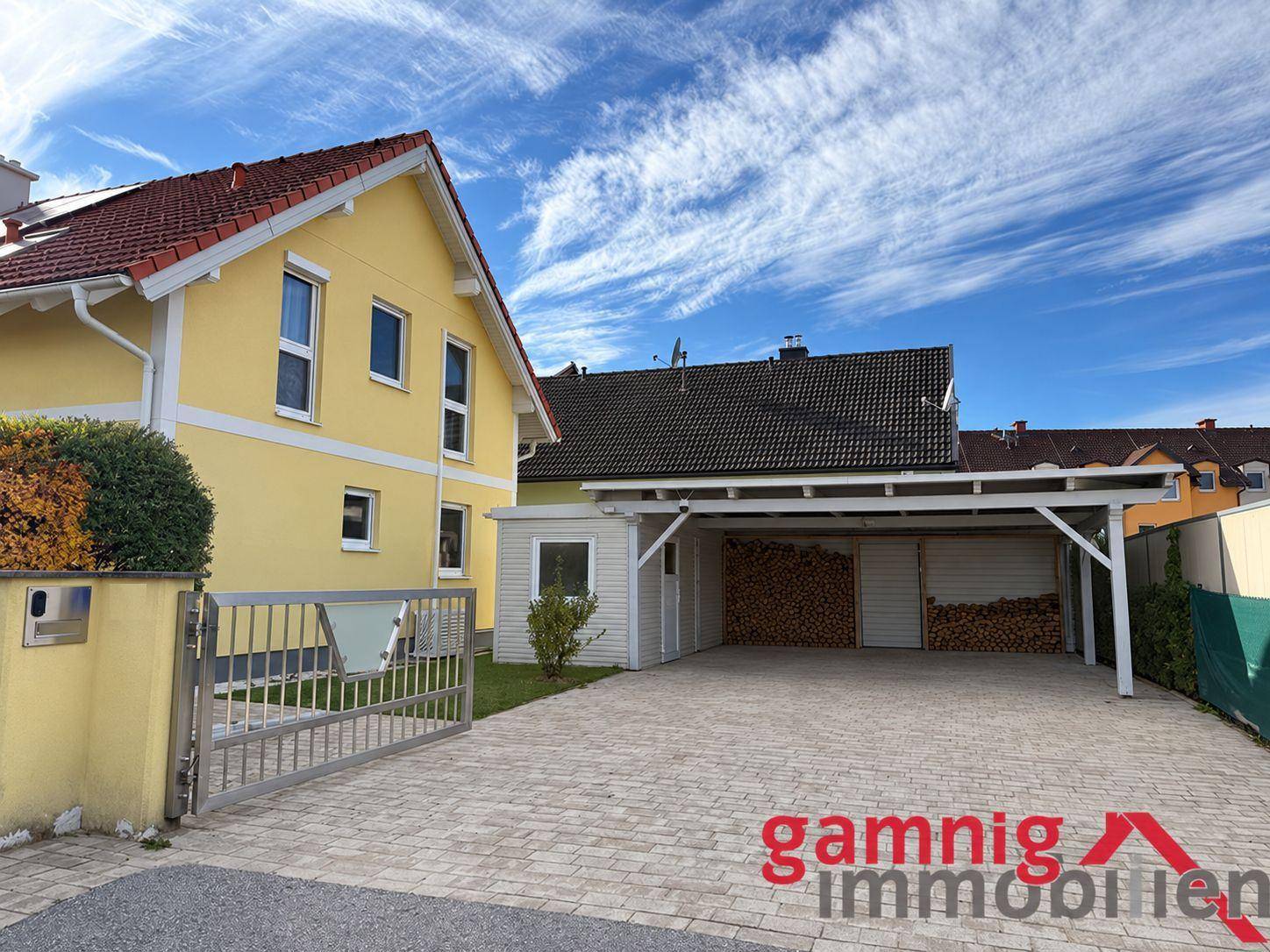 Einfahrt mit Carport und Holzstapel neben dem gelben Einfamilienhaus.