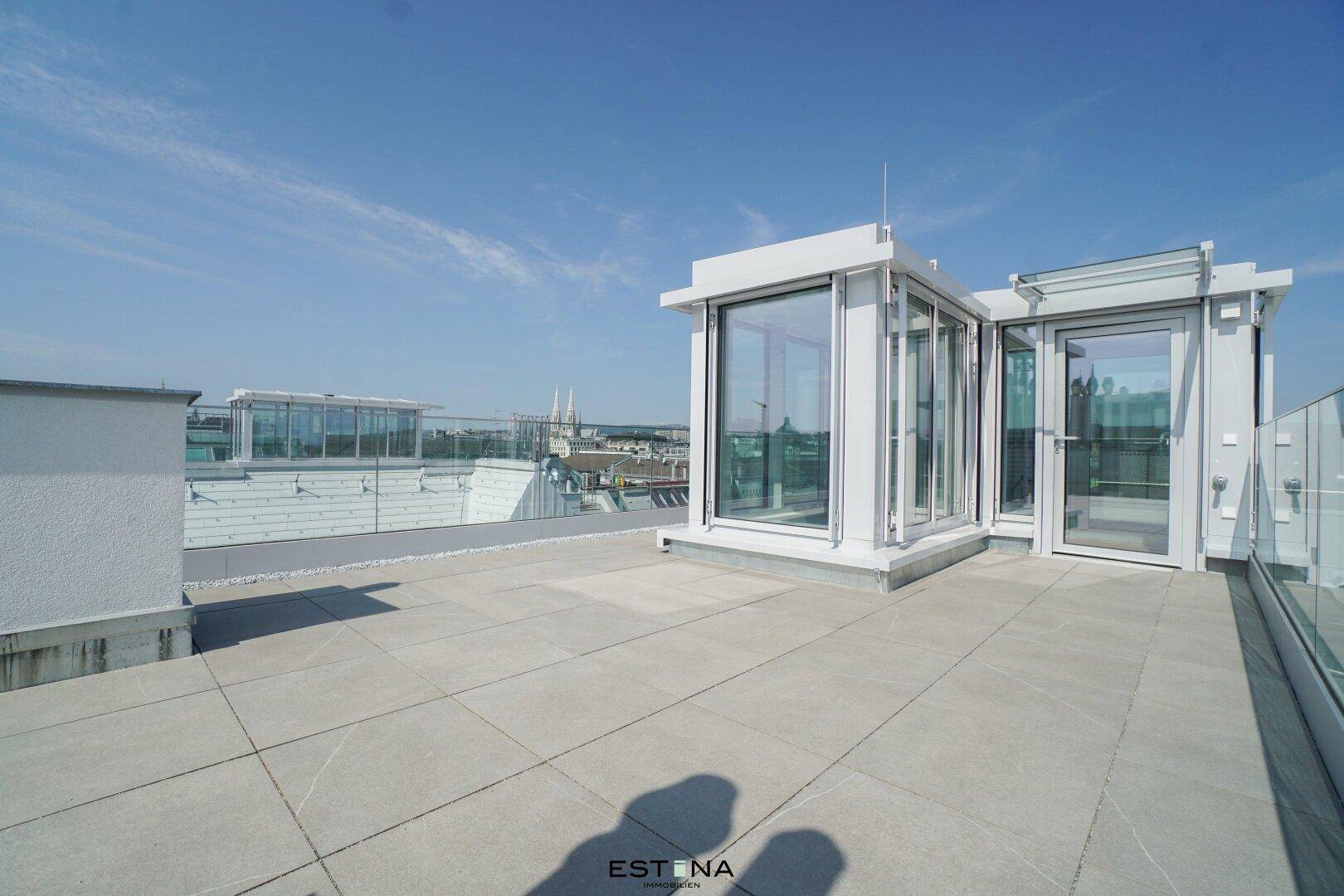 Geräumige Dachterrasse mit Glasgeländer und Zugang zu einem verglasten Dachaufbau mit Stadtblick.