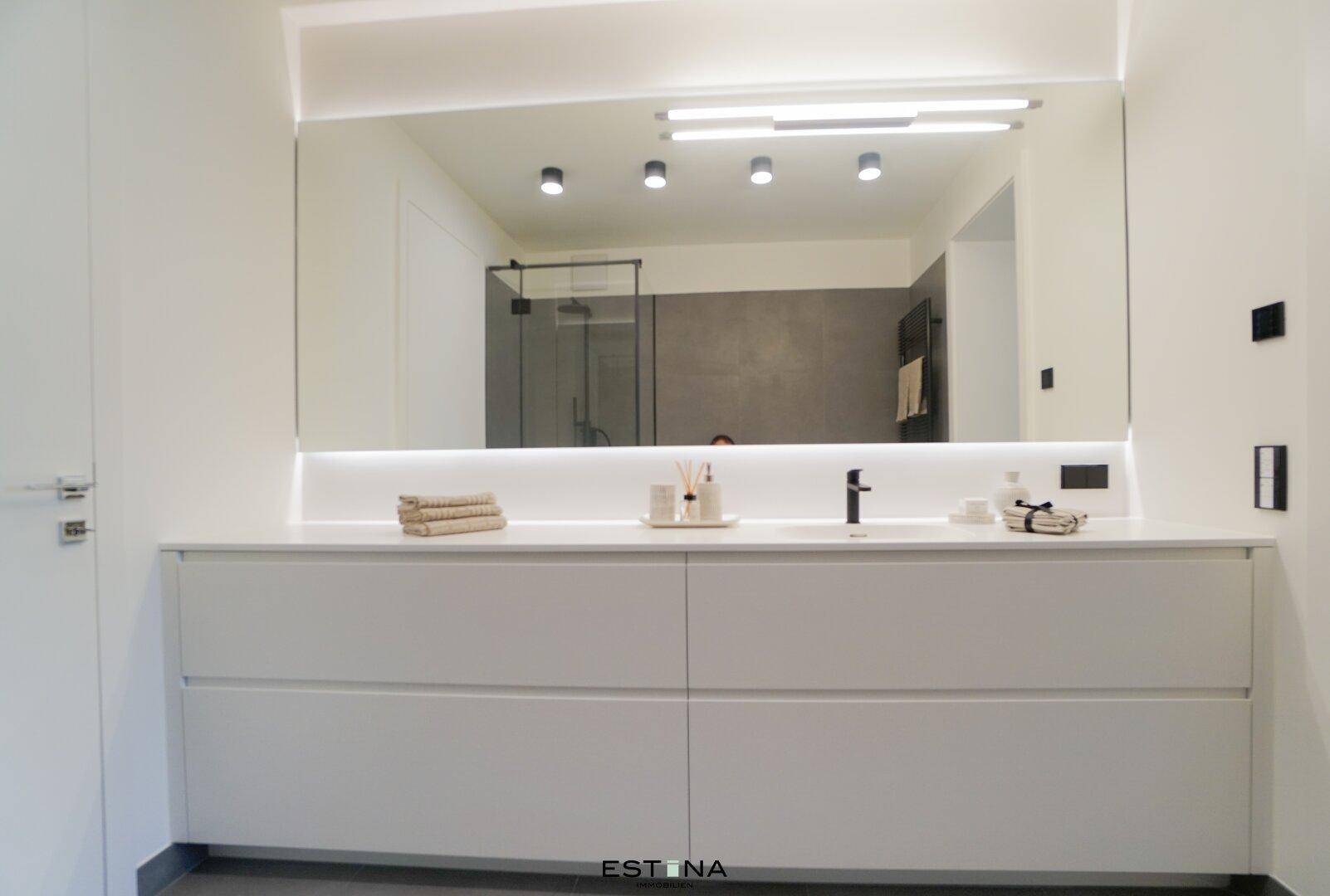 Modernes Badezimmer mit Doppelwaschbecken, großem beleuchtetem Spiegel und einer begehbaren Dusche im Hintergrund.