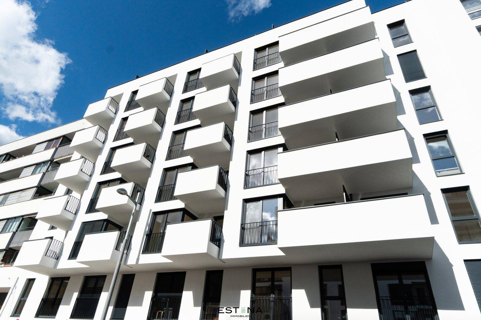 Moderne Wohnhausfassade mit vielen Balkonen und großen Fenstern unter blauem Himmel.