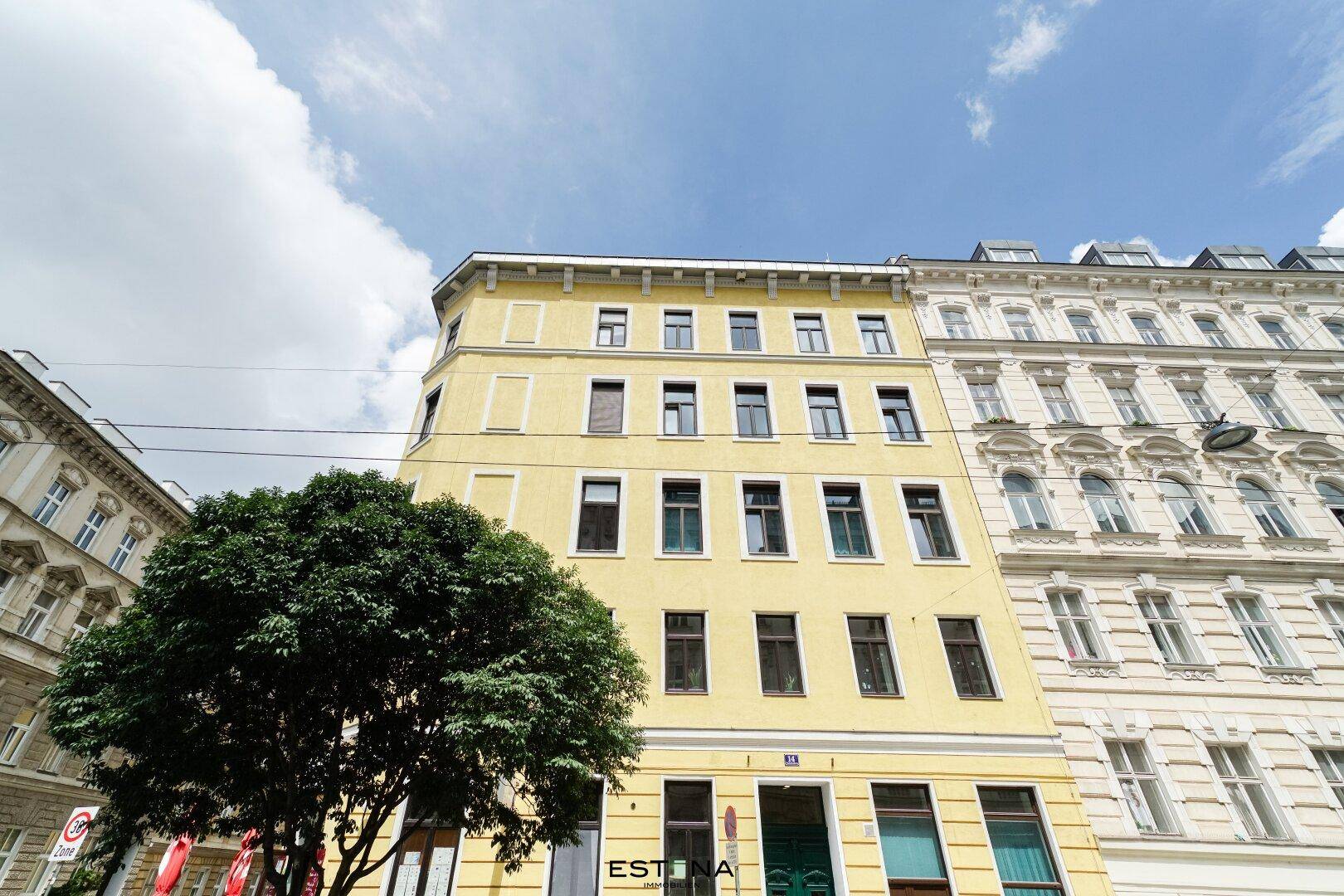 Historisches Gebäude mit gelber Fassade und vielen Fenstern unter blauem Himmel, umgeben von Stadtbäumen.