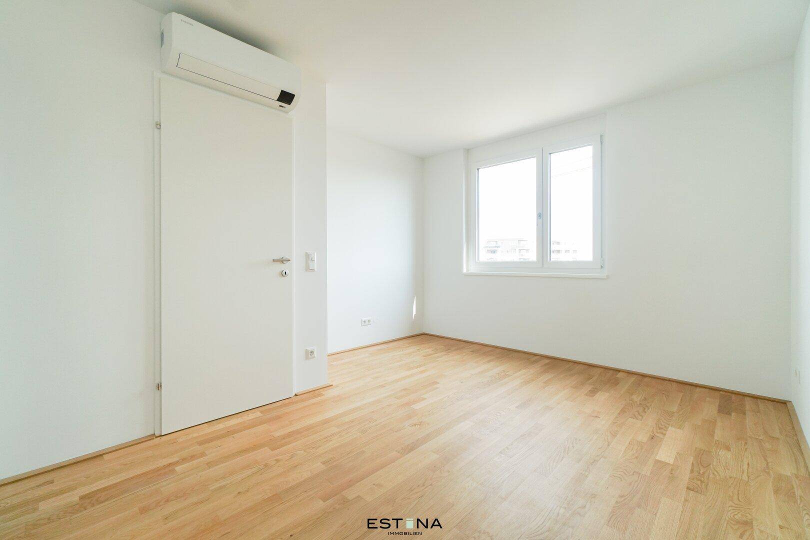 Helles, leeres Zimmer mit Holzboden, weißen Wänden und Klimaanlage, ideal als Schlafzimmer.