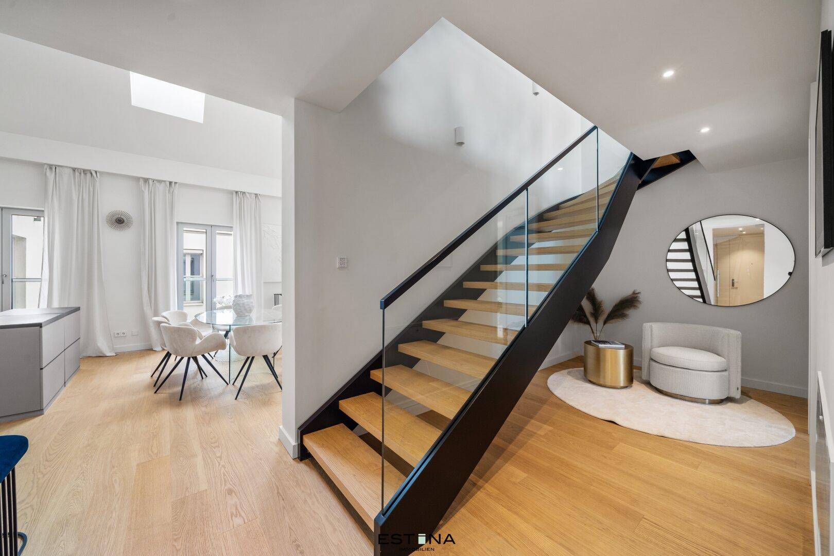 Heller Flur mit moderner Holztreppe, Glasgeländer und Blick auf den Wohnbereich.