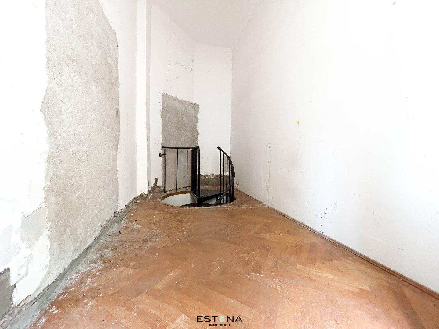 Blick von oben auf eine schwarze Wendeltreppe in einem Raum mit renovierungsbedürftigem Holzboden.