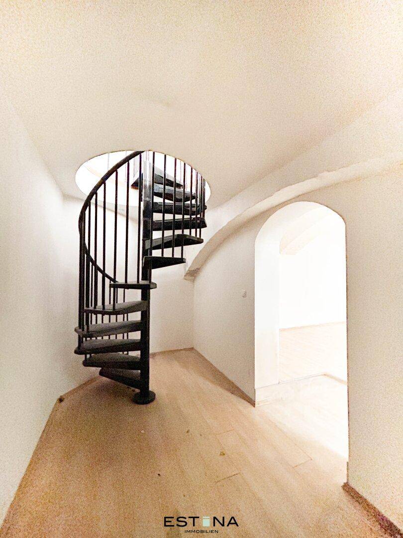 Elegante schwarze Wendeltreppe in einem hellen Flur mit gewölbtem Durchgang und Holzboden.