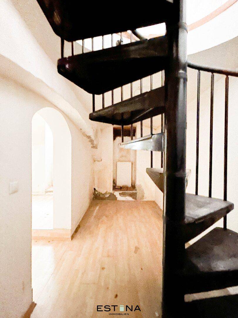 Dunkle Wendeltreppe in einem schmalen Flur mit hellem Holzboden und gewölbten Durchgängen.