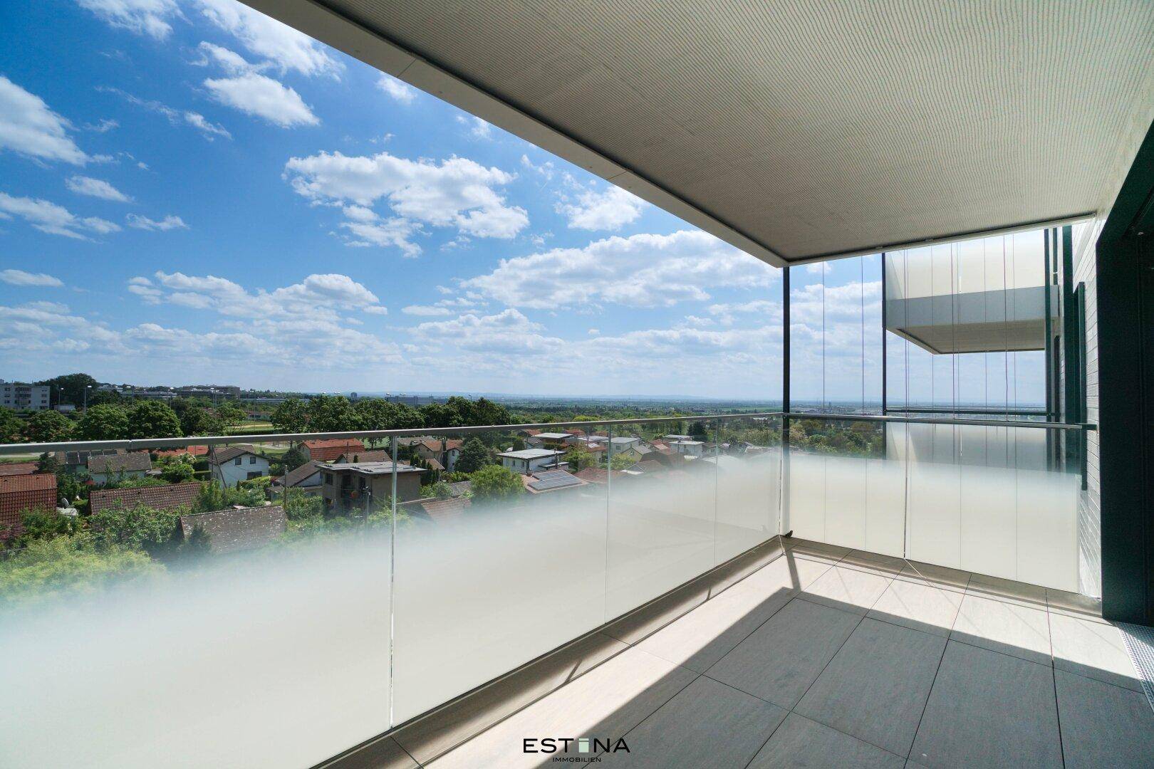 Moderner Balkon mit Glasgeländer und weitem Blick über die grüne Wohngegend unter blauem Himmel.