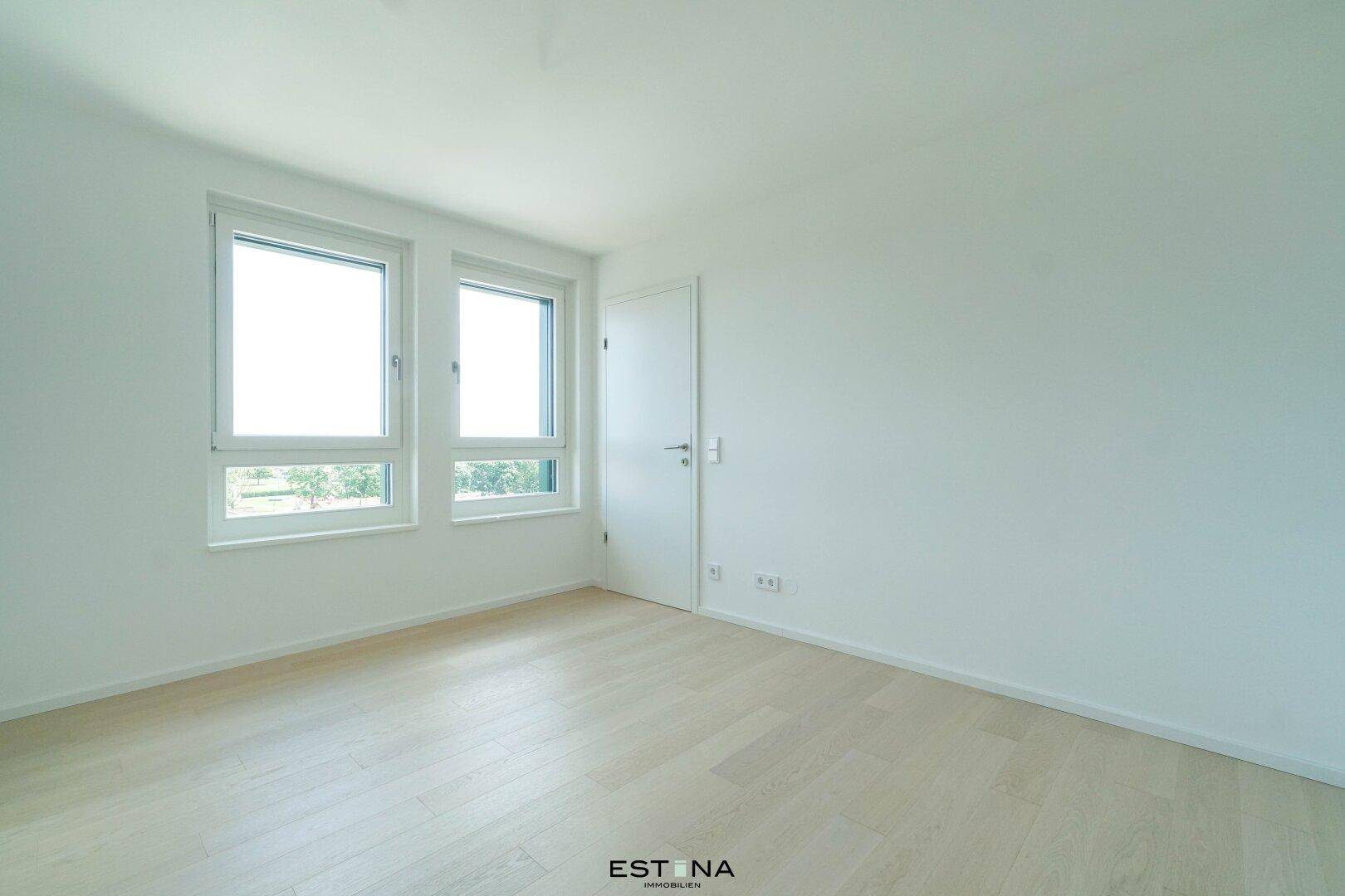 Leerer Raum mit hellen Wänden, Parkettboden und zwei Fenstern, die viel Tageslicht hereinlassen.