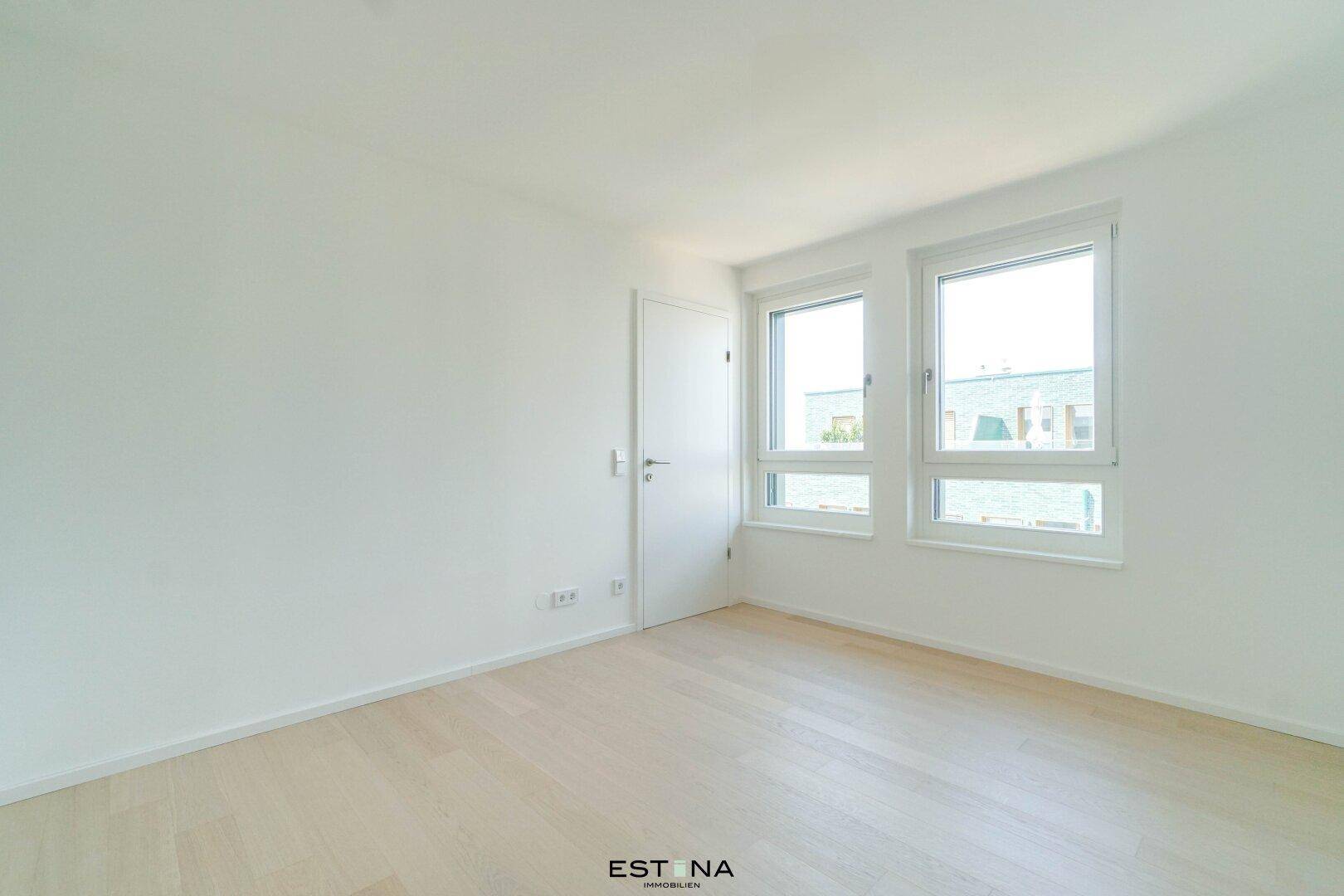 Helles Zimmer mit zwei Fenstern, hellem Parkettboden und weißen Wänden, ideal als Schlafzimmer.