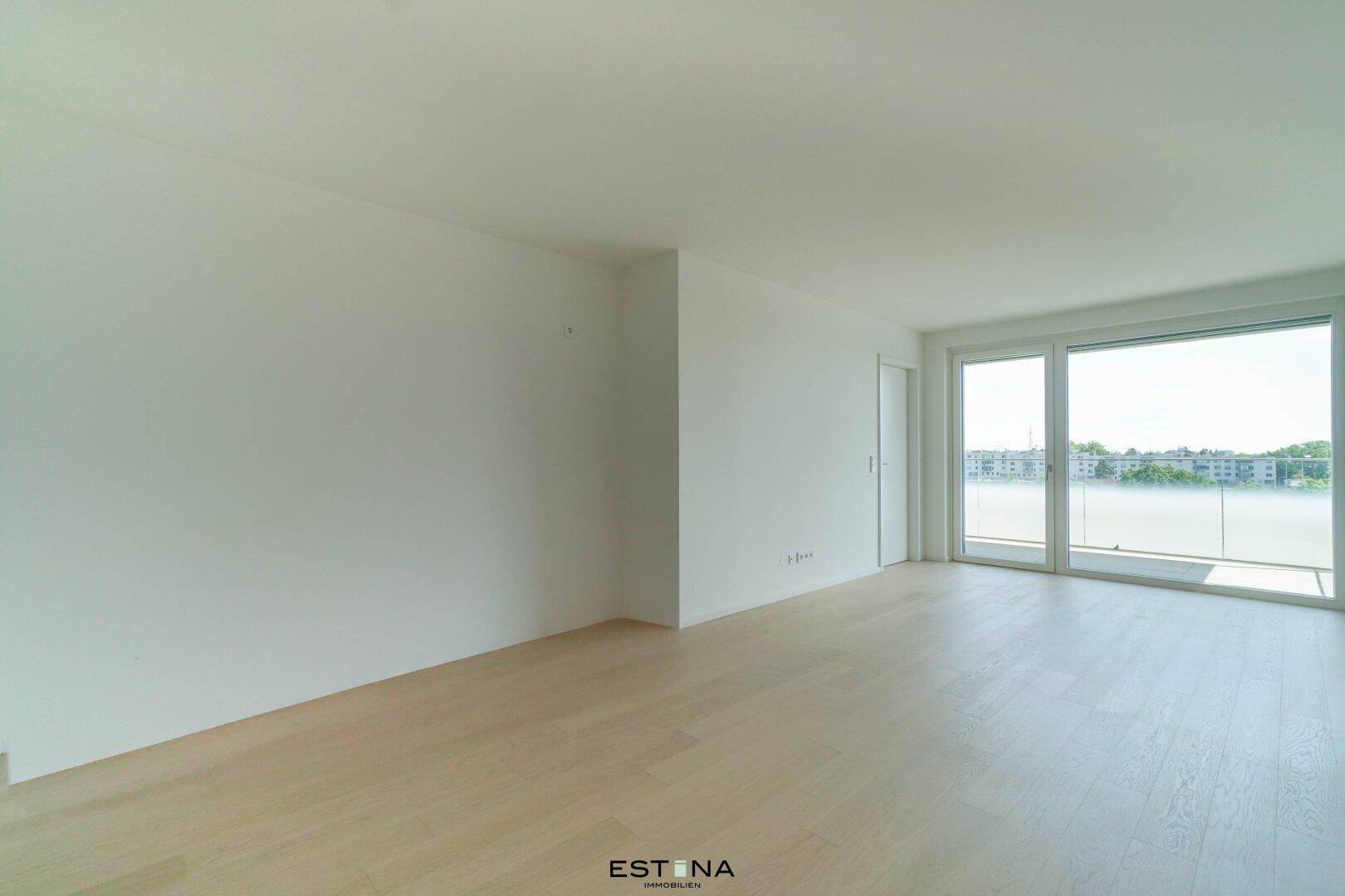Helles, leeres Wohnzimmer mit hellem Holzfußboden und großen Fenstern zum Balkon.