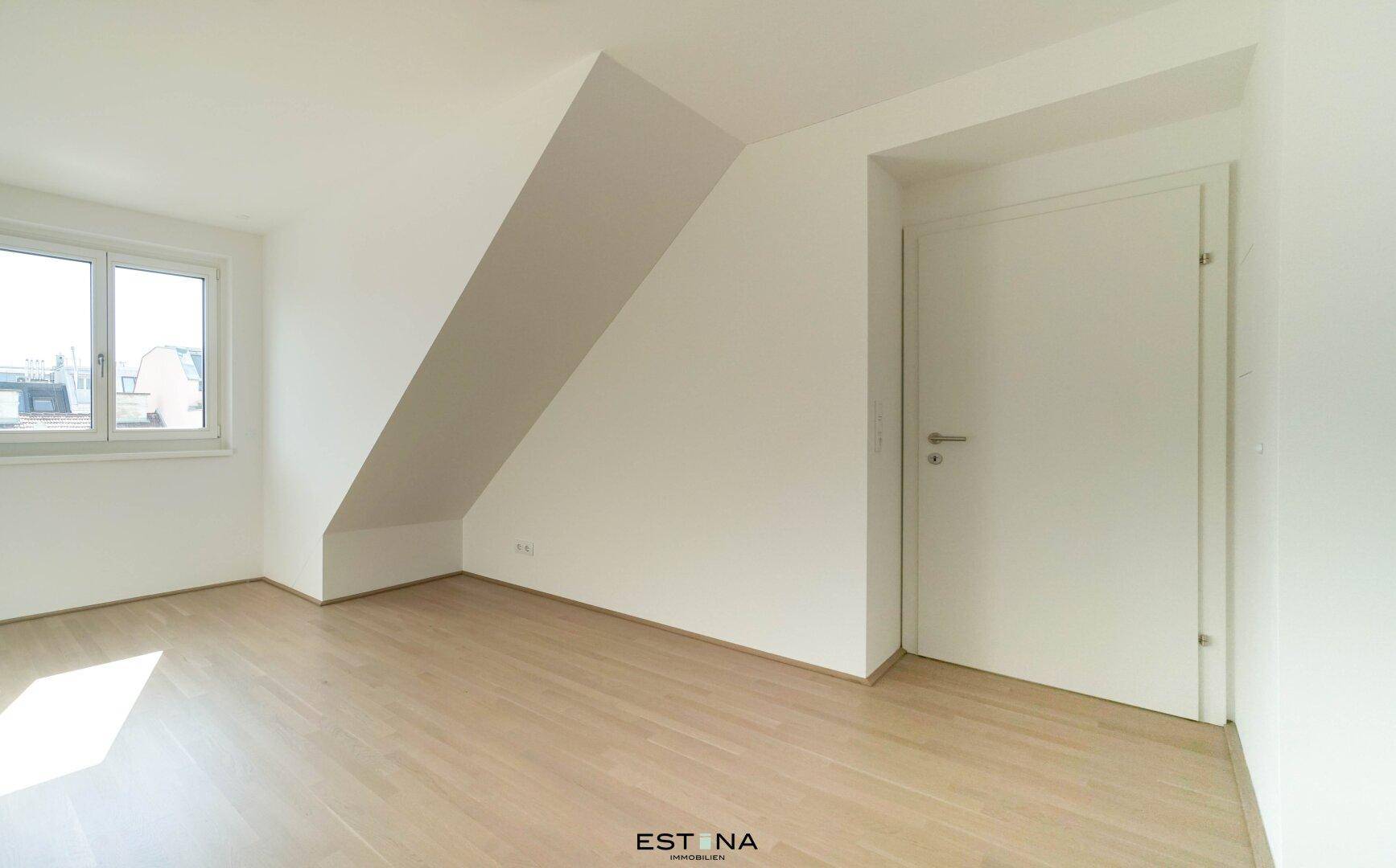 Leerer Raum in Dachgeschosswohnung mit Holzboden, weißer Tür und einem Fenster.