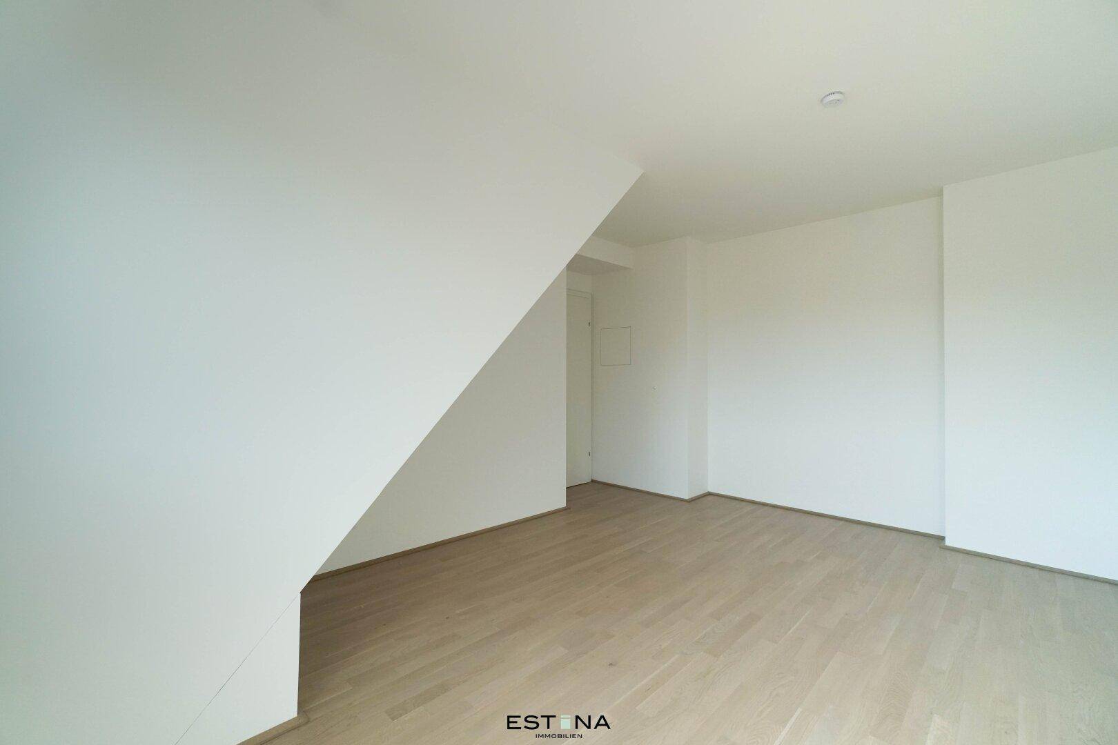 Geräumiger, unmöblierter Raum in Dachgeschosswohnung mit schrägen Wänden und hellem Holzboden.