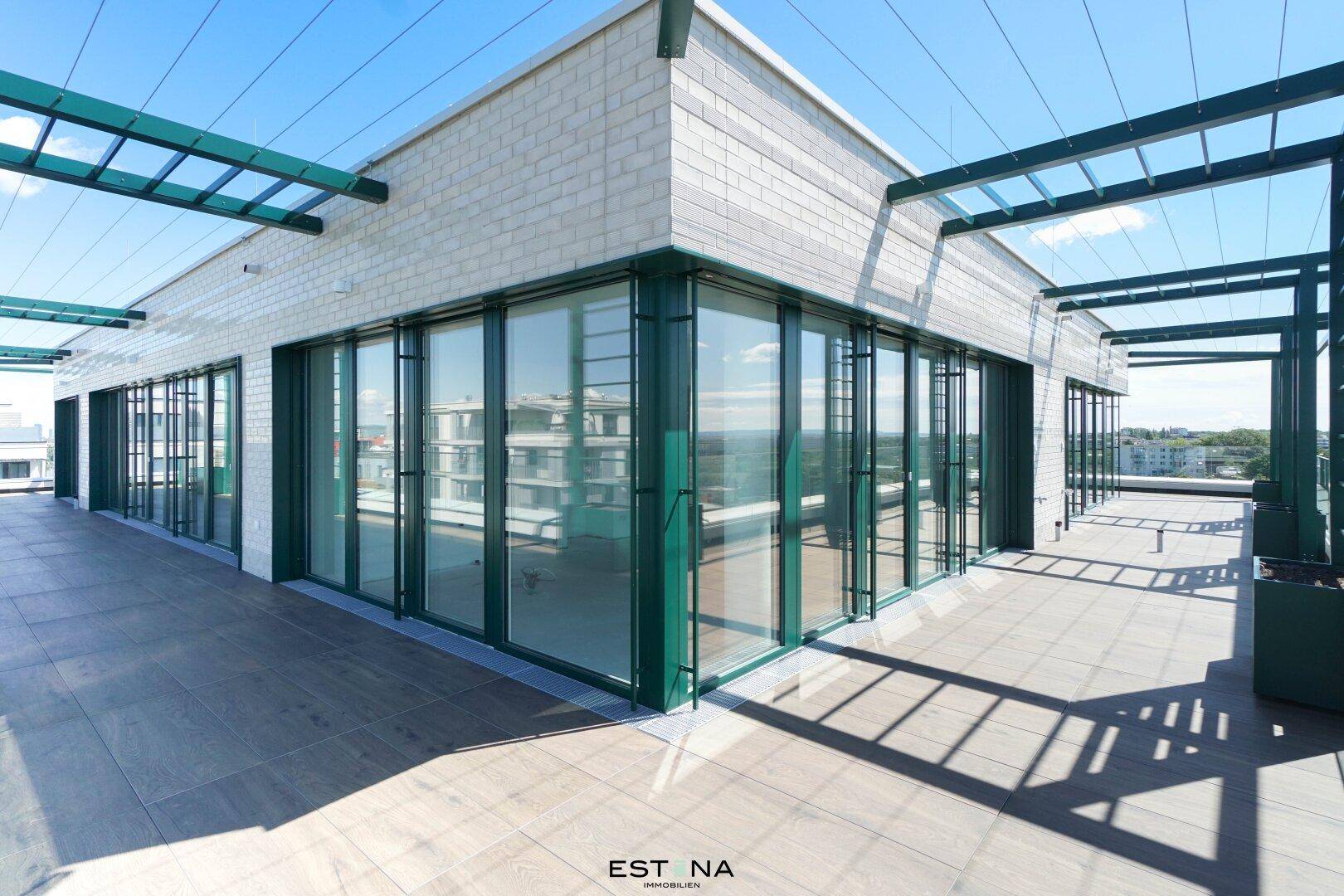 Moderne Gebäudeecke mit großer Terrasse, Glasgeländer und grünen Metallstrukturen.