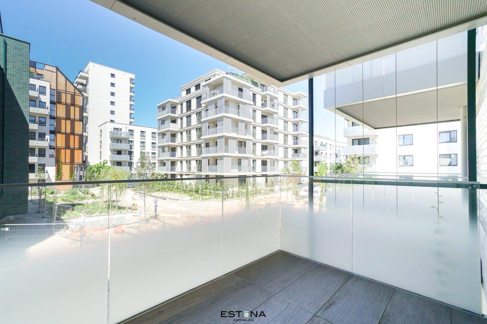 Balkon mit Glasgeländer und weitem Blick auf moderne Gebäude und eine grüne Umgebung.