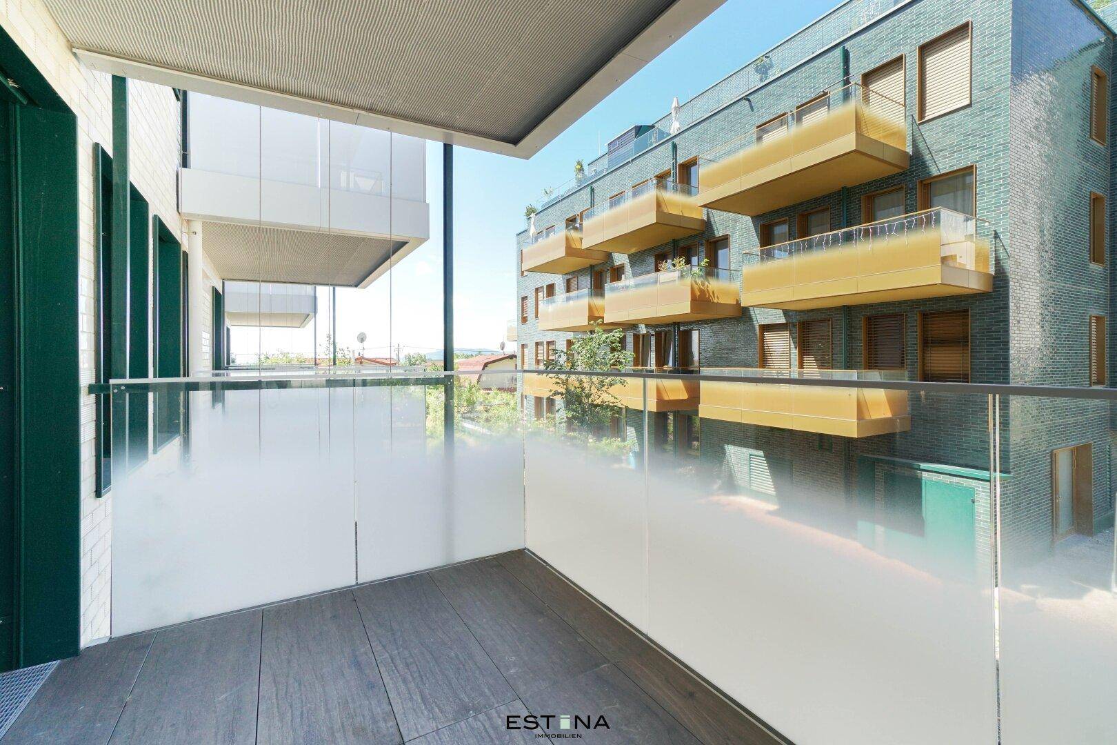 Moderner Balkon mit Glasgeländer und Blick auf umliegende Wohngebäude unter blauem Himmel.