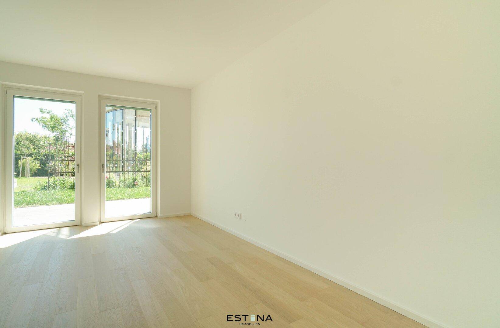 Leeres Zimmer mit hellem Parkettboden und zwei großen Fenstertüren zum Garten.