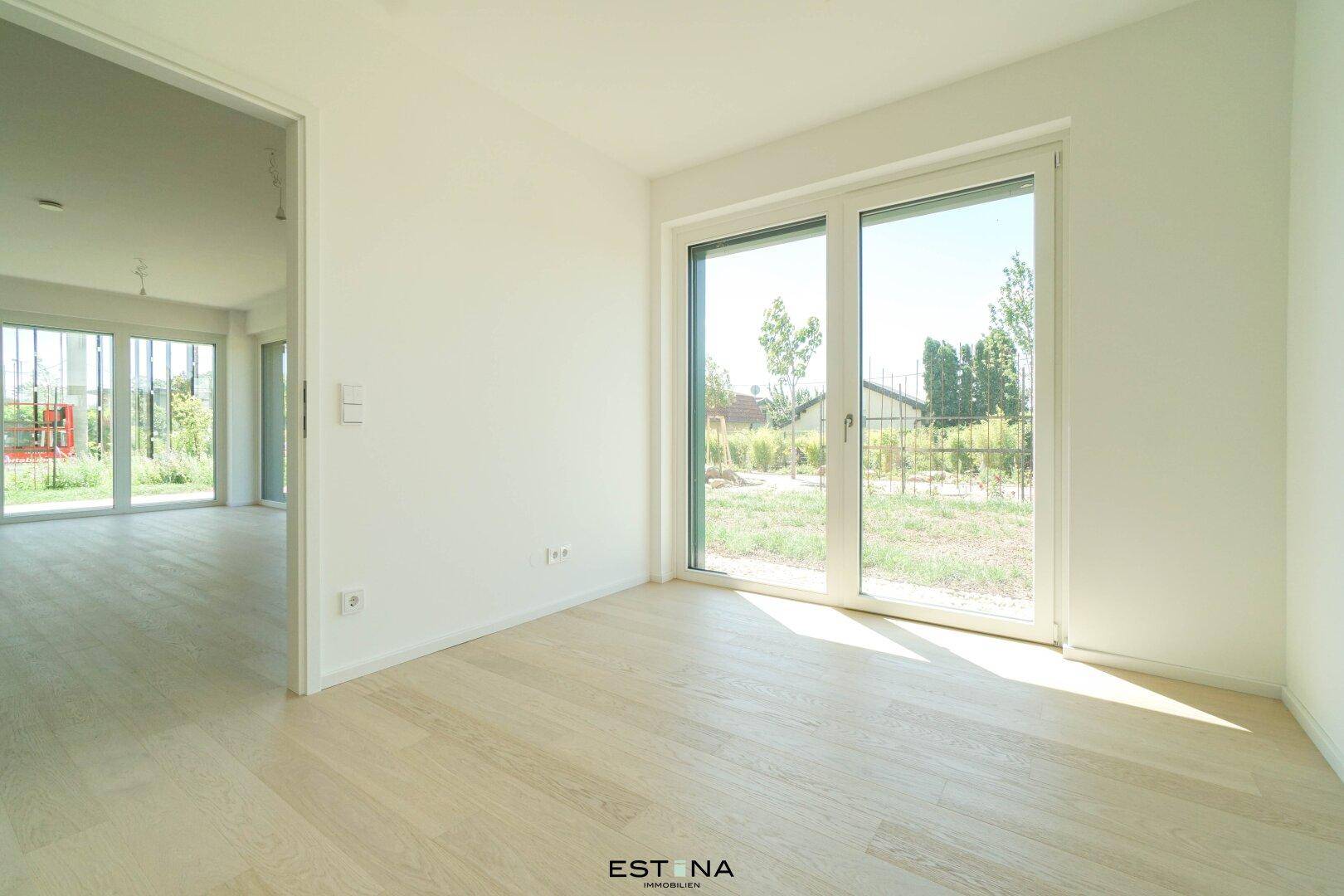 Helles Schlafzimmer mit großen Fenstern, hellem Parkettboden und Zugang zu einem weiteren Raum.