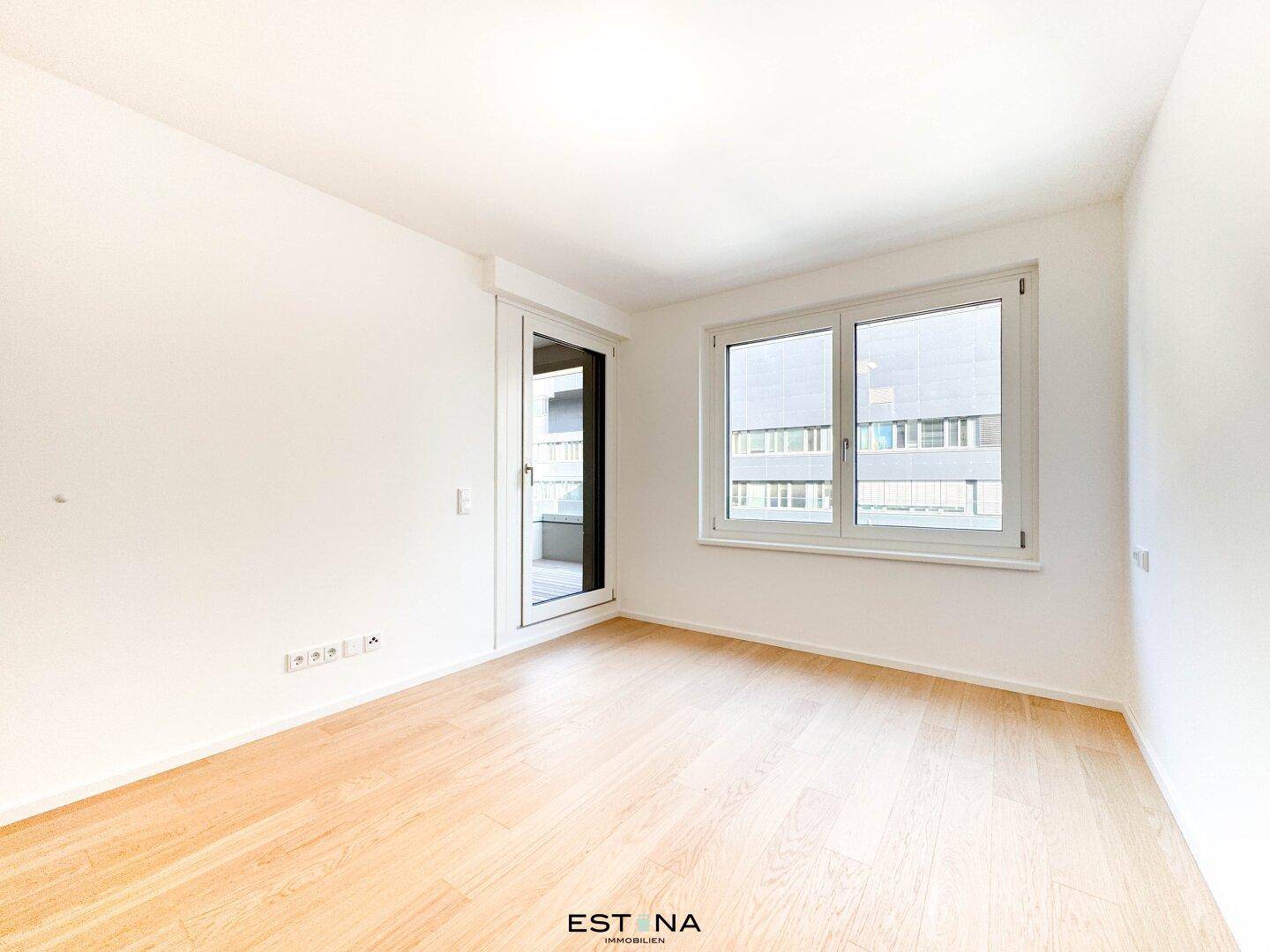 Helles, leeres Zimmer mit Parkettboden und Zugang zu einem Balkon oder einer Loggia.